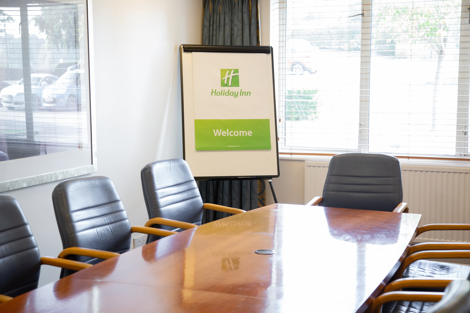 A conference room with a wooden table surrounded by black leather chairs. A flip chart with a Holiday Inn welcome sign is near the window, which has blinds and curtains. Outside, cars are visible through the window.