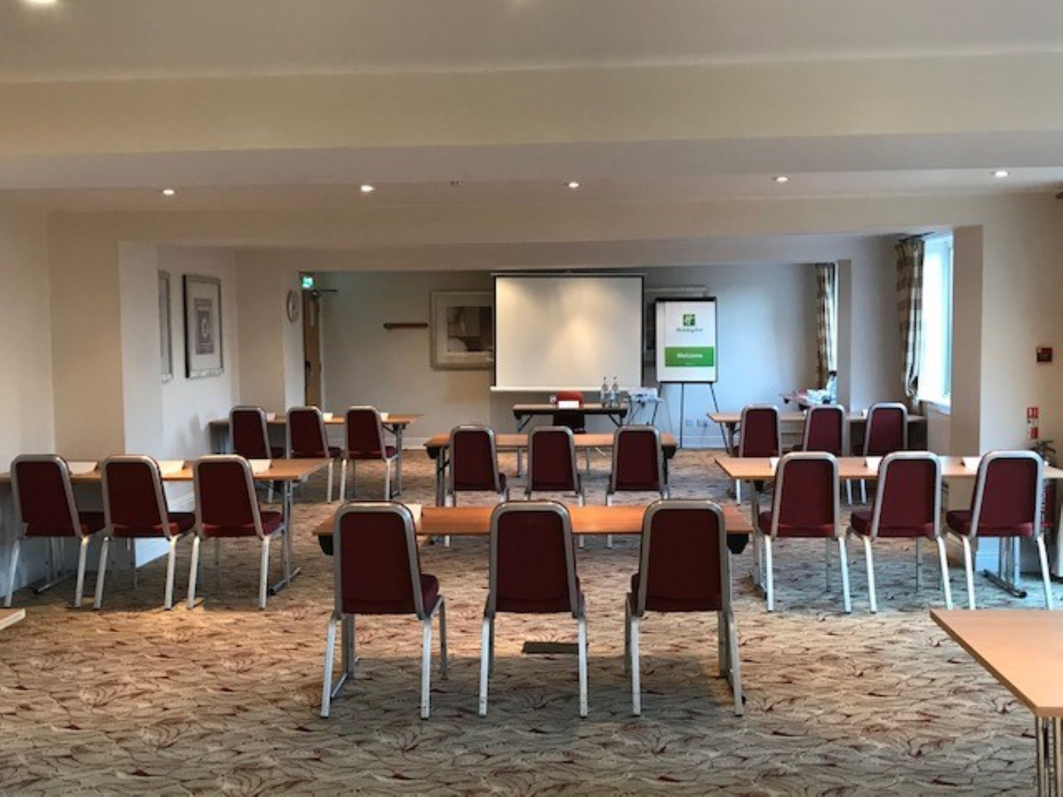 Empty conference room with multiple rows of chairs and tables, a whiteboard, a projection screen, and a projector, set up for a presentation.