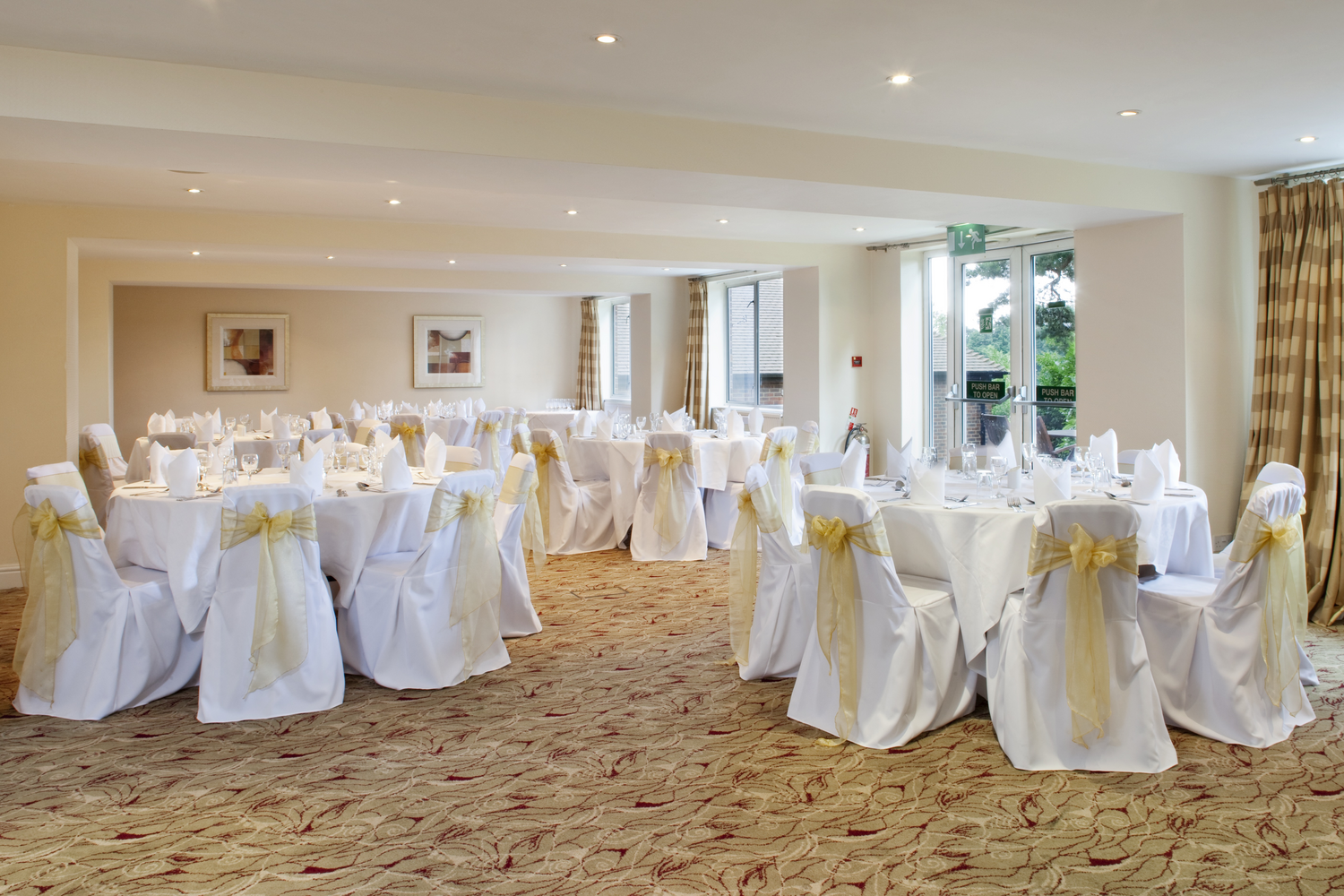 Elegant banquet hall decorated for a formal event with white tablecloths and chairs with yellow bows, set with glassware and napkins.