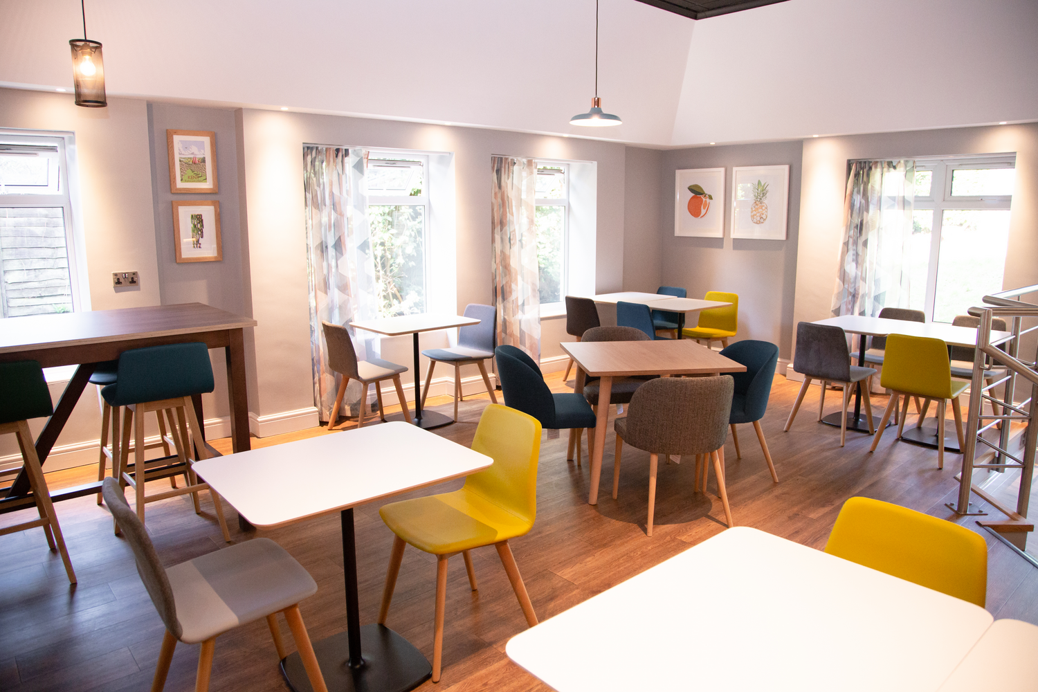 Bright, modern cafe with white walls, large windows, colorful chairs, and wooden tables. Decor includes framed artwork of fruits and plants, with curtains featuring geometric patterns. Overhead lighting provides warm illumination.