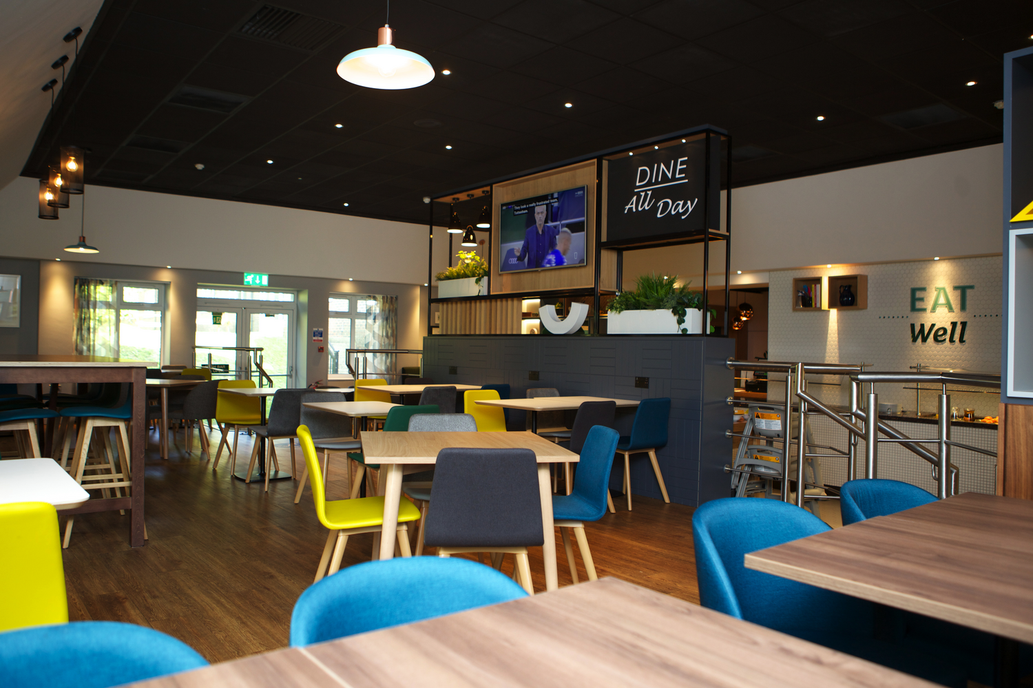 Interior of a modern restaurant or café with colorful chairs and wooden tables, a television on the wall, and signs that say "DINE All Day" and "EAT Well."