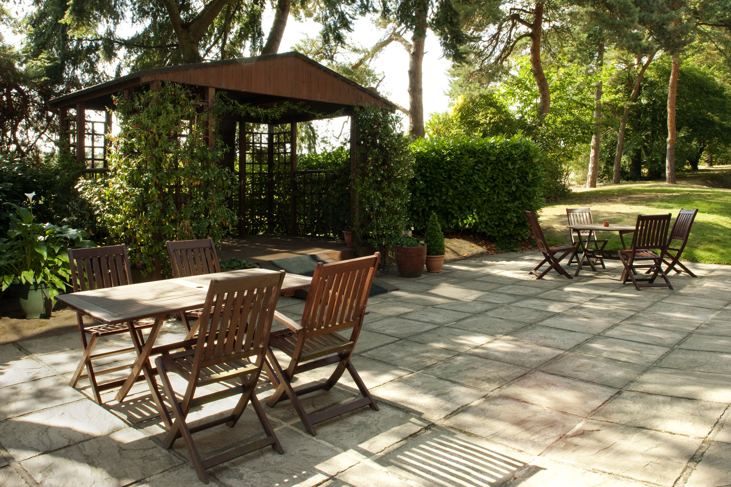 Outdoor patio with wooden tables and chairs, surrounded by lush green trees and plants, with a wooden gazebo in the background on a sunny day.