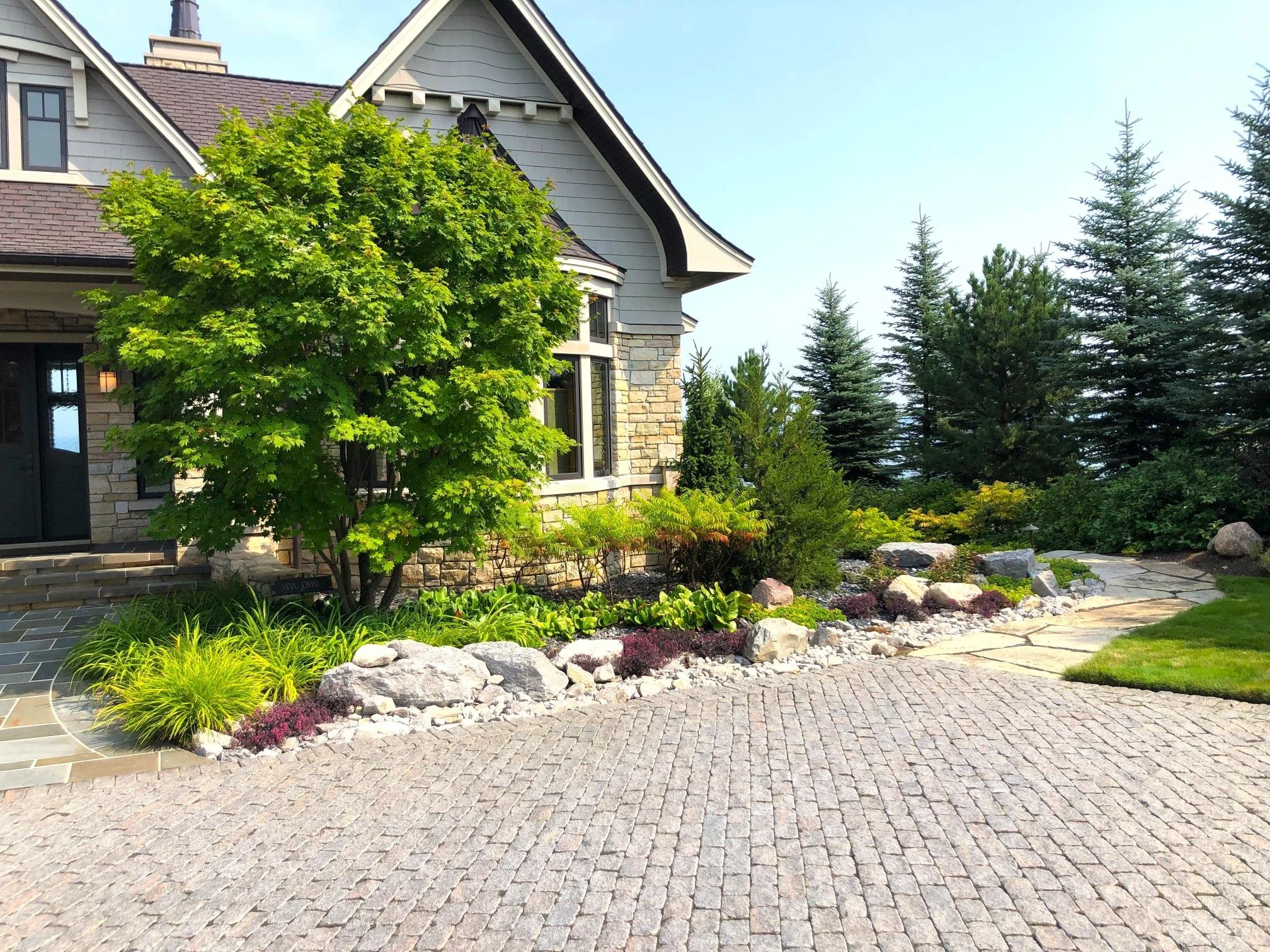 A residential house with a stone and shingle exterior, surrounded by a landscaped garden with various trees, shrubs, and rocks, and a patterned stone pathway in the foreground.
