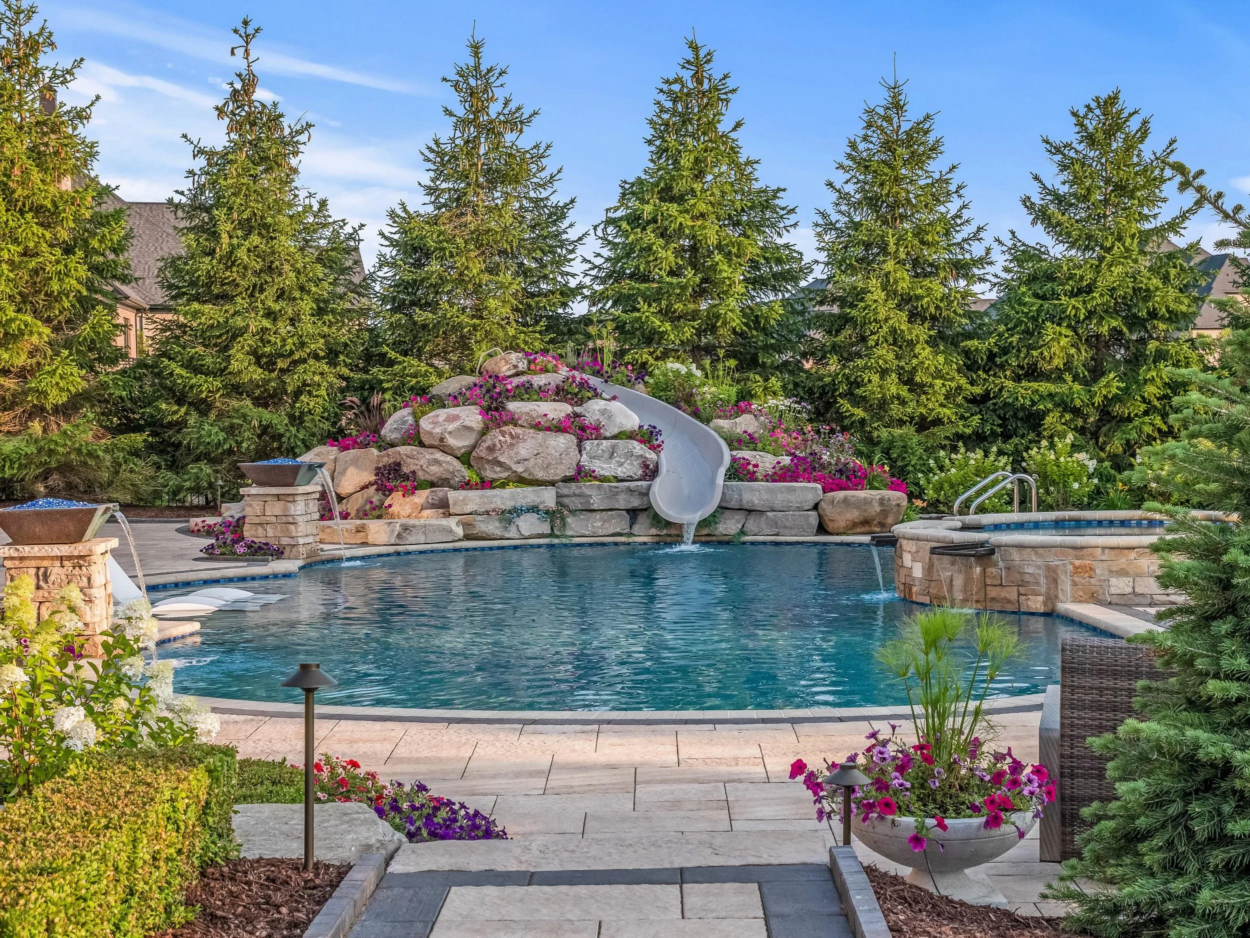A backyard pool area with a fire pit, lush greenery, and decorative flowers, surrounded by trees and houses, on a clear day.