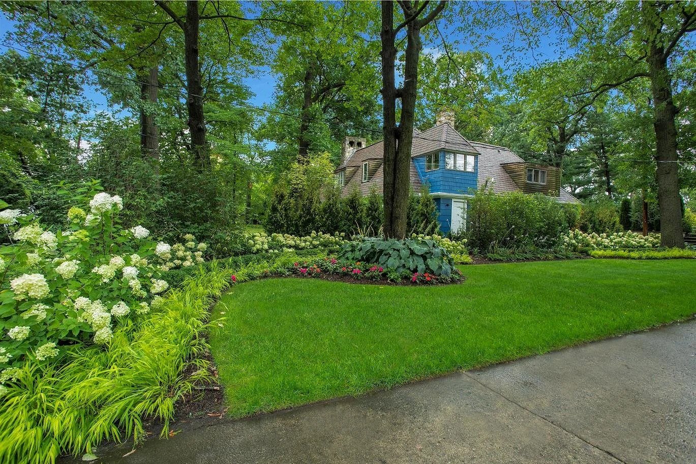 A well-maintained front yard with green grass, decorative shrubs, and blooming flowers, in front of a blue and brown house surrounded by tall trees.