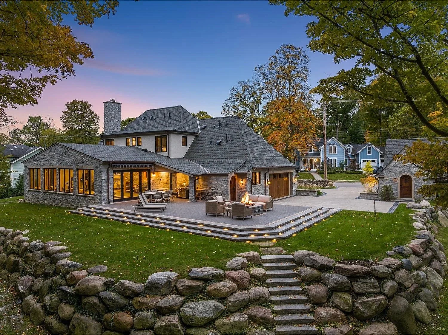 A large, two-story house with a gray stone exterior and shingled roof, illuminated from within, with a backyard patio at dusk. The patio has a fire pit, outdoor furniture, and lighting on the steps, surrounded by a stone retaining wall, with trees and neighboring houses visible in the background.