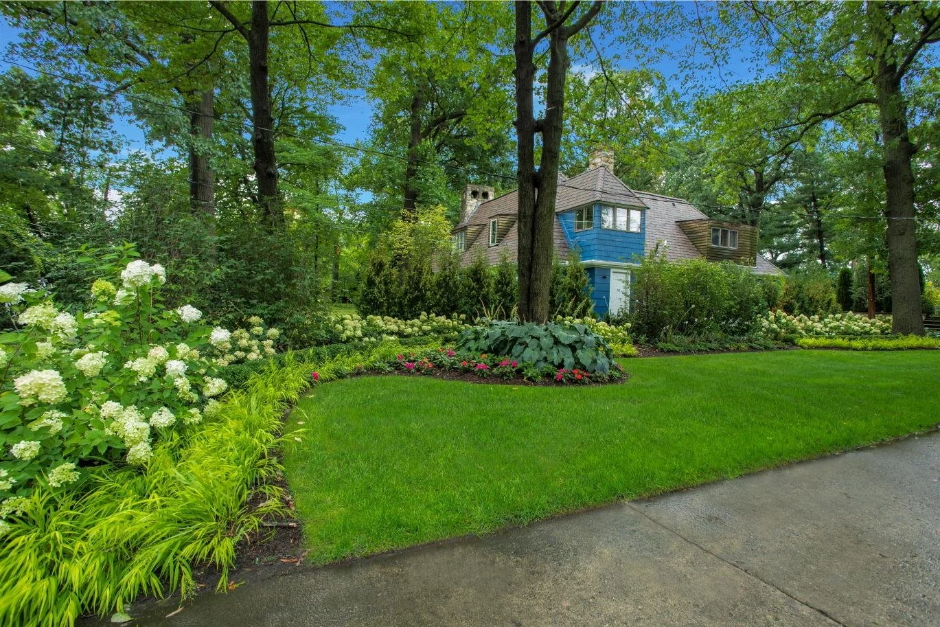A well-maintained front yard with green grass, colorful flower beds with white, pink, and green plants, surrounding a charming house hidden among tall trees with lush green foliage, in a sunny day.