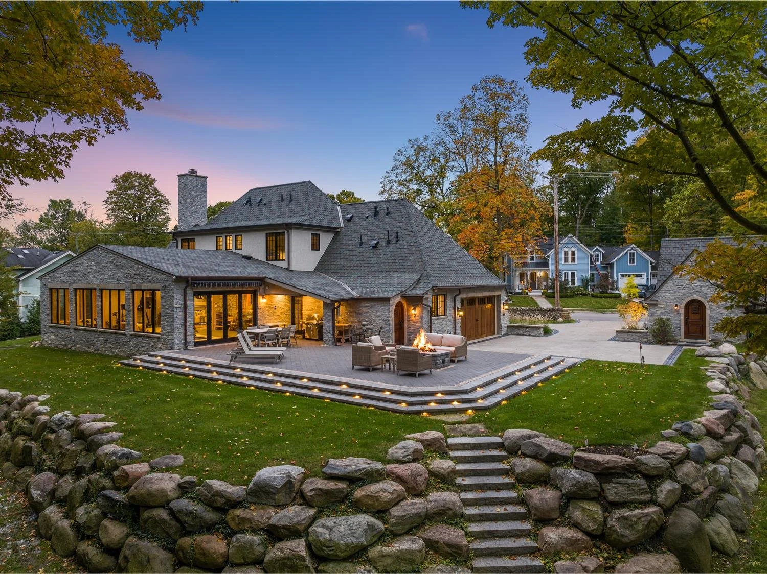 A large house with a stone and stucco exterior, multiple rooflines, and large windows glowing from interior lighting. The house features an outdoor patio with a fire pit, seating, and stairs leading down to a grassy yard enclosed by a stone wall. Surrounding trees have fall foliage, and neighboring houses are visible in the background during dusk.