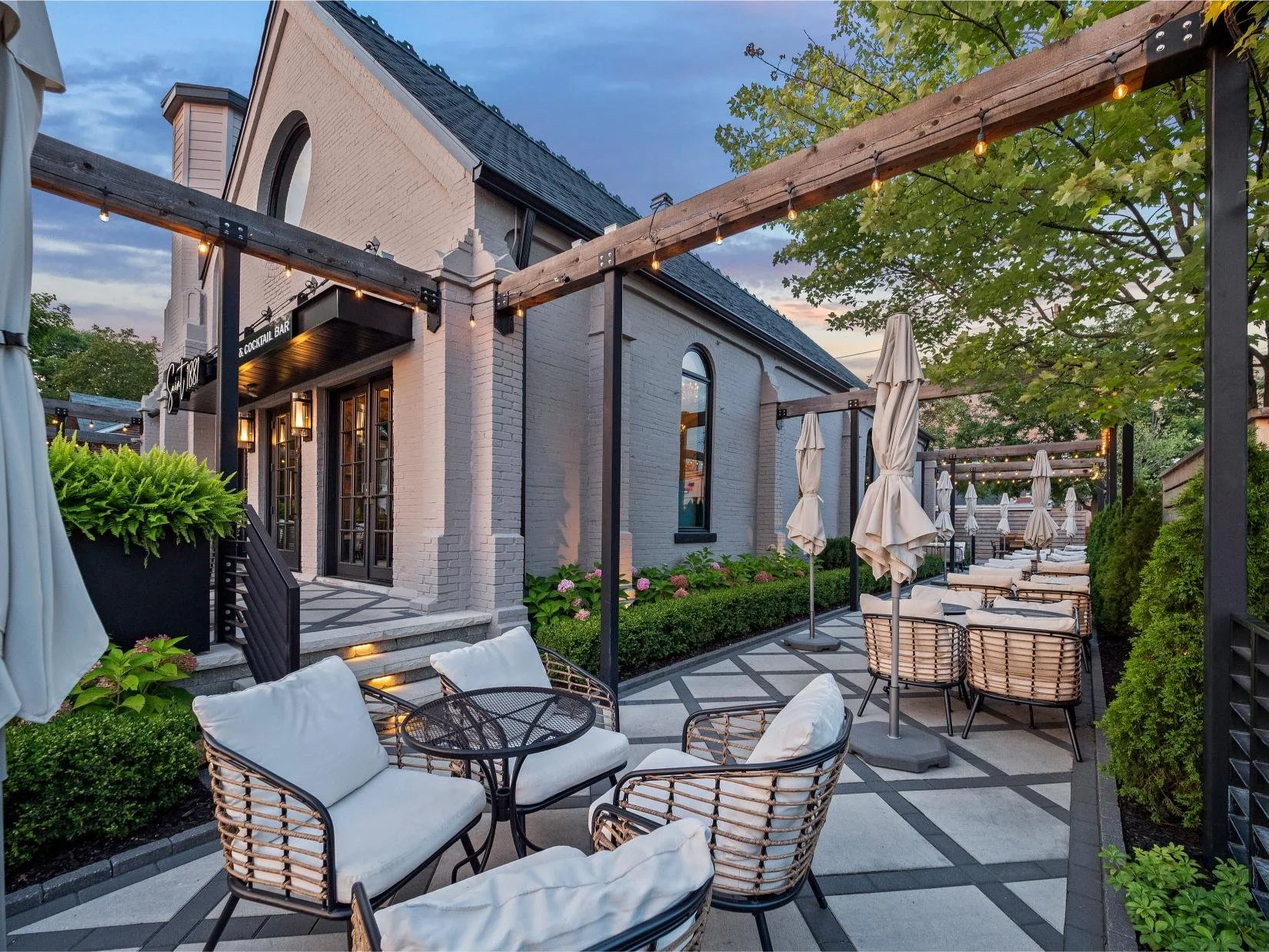 Outdoor patio area with cushioned chairs, umbrellas, and string lights, adjacent to a brick building with black-framed windows and a sign that reads 'Cocktail Bar' at dusk.