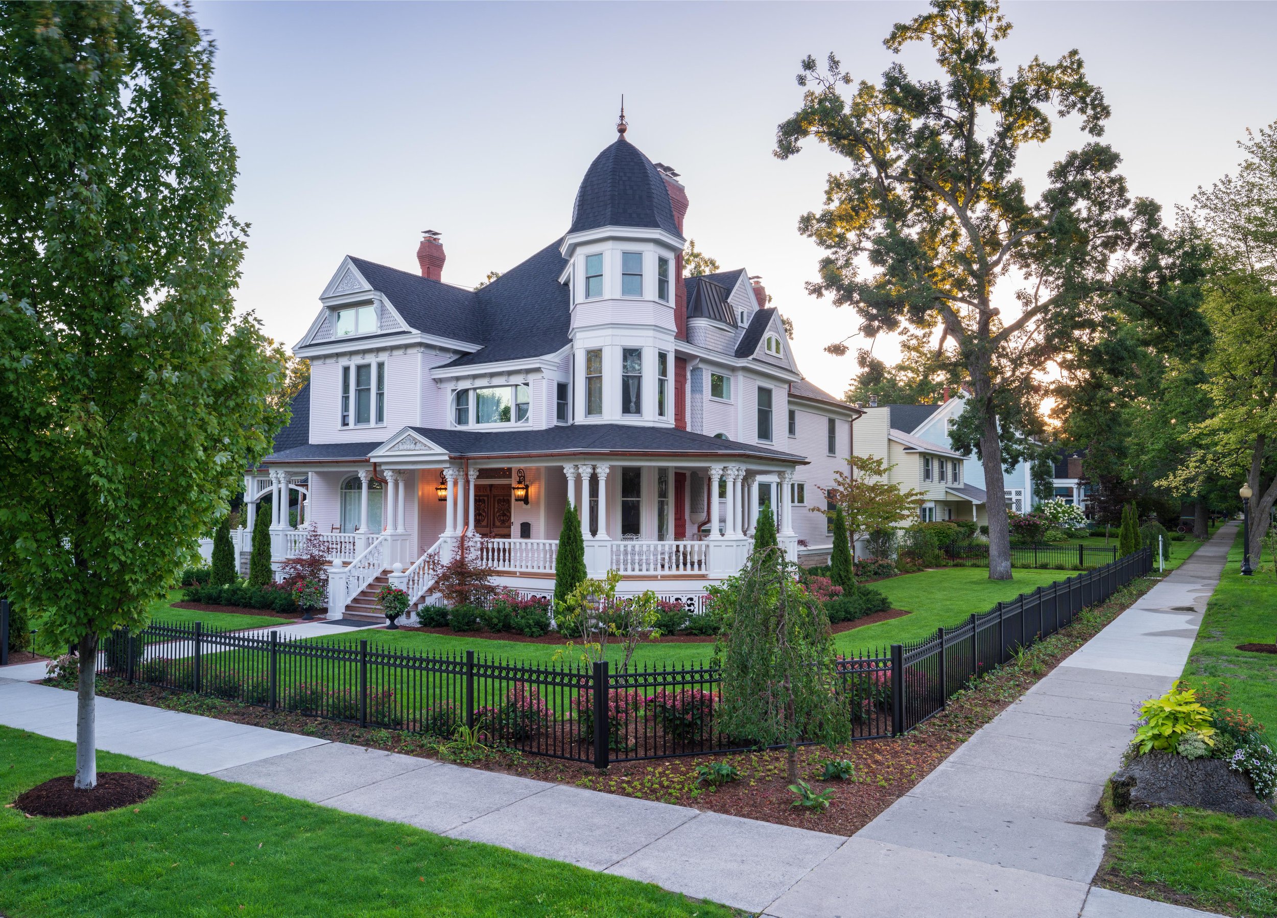 A Victorian-style house with a turret, white siding, and a front porch, surrounded by a black picket fence and landscaped garden with green grass, trees, and flowering plants, during daylight.