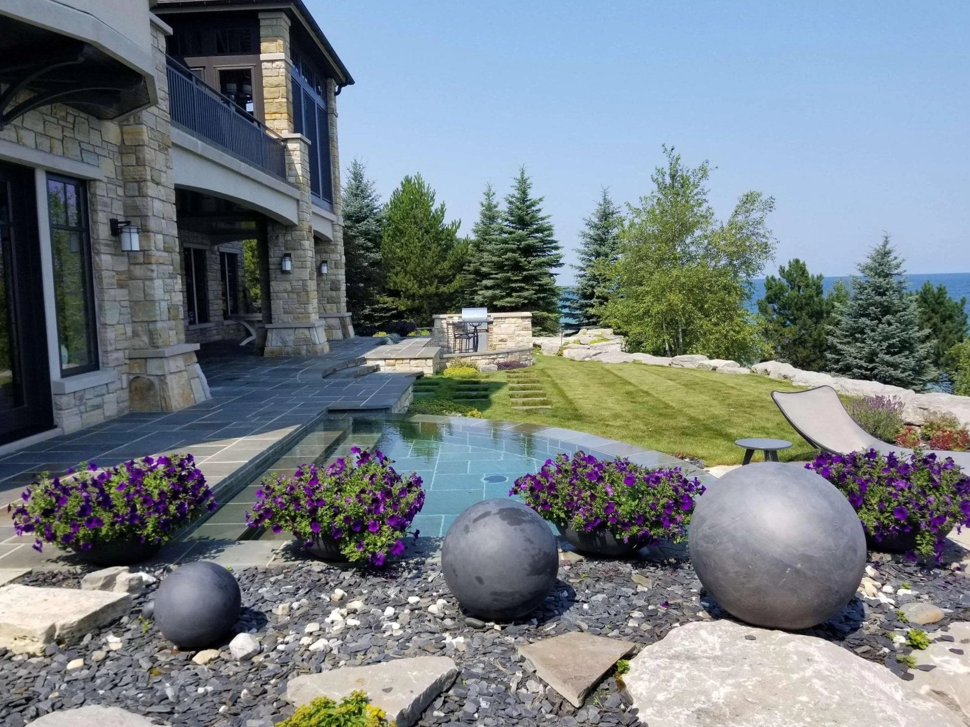 An outdoor patio area features a stone house with a balcony, a neatly mowed lawn, a fire pit, and a garden with purple flowers. There are large round stones in the garden and some trees in the background, with blue sky and ocean visible beyond the trees.