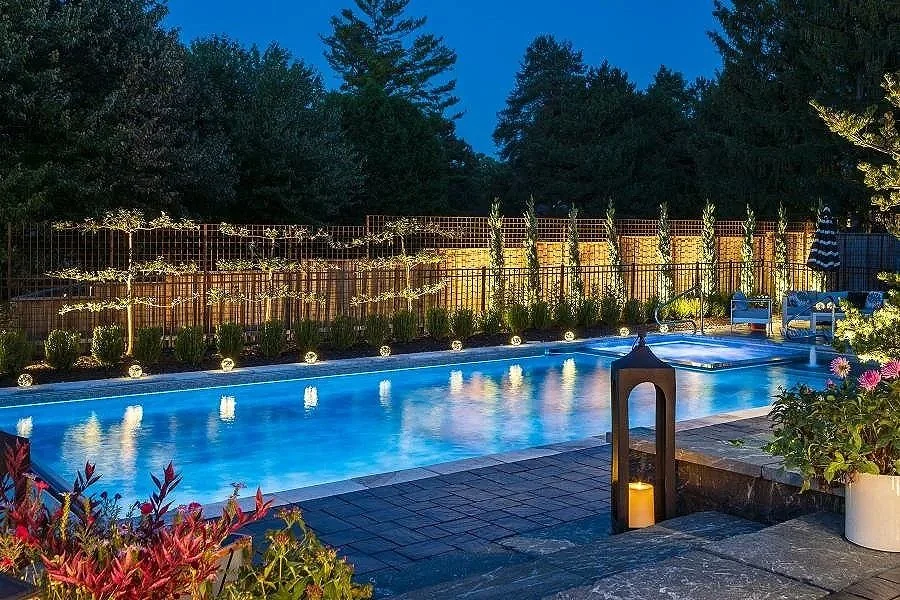Backyard swimming pool at dusk, illuminated by string lights and small lights along the fence, with plants and flowers around and trees in the background.