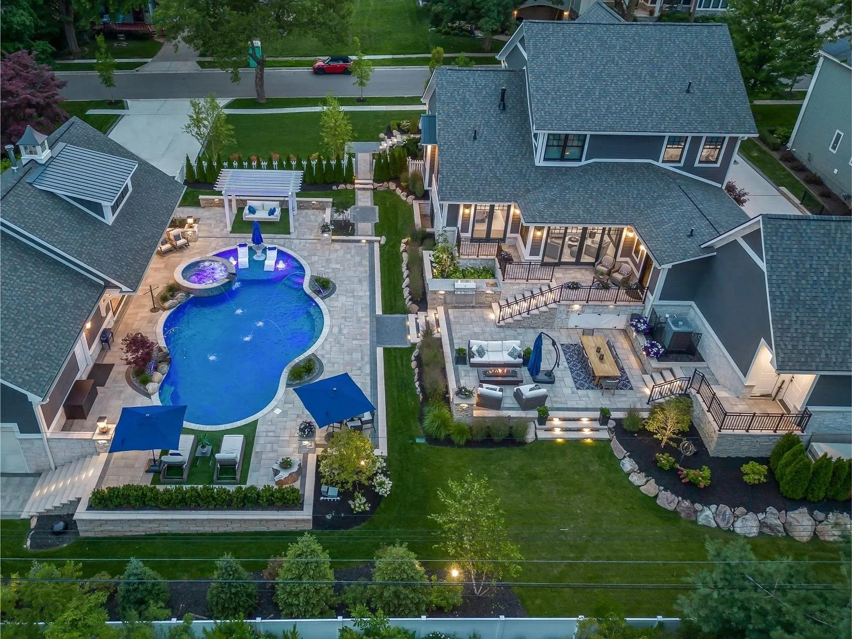 Aerial view of a modern backyard at dusk featuring a kidney-shaped swimming pool with fountains, a hot tub, and multiple patio areas with outdoor furniture and umbrellas, surrounded by well-kept grass, trees, and landscaped garden beds.