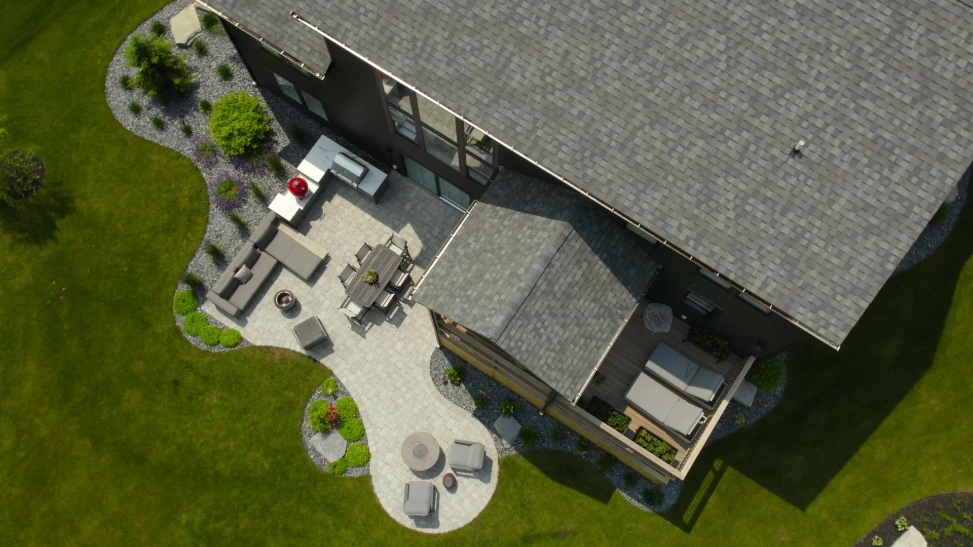 Aerial view of a house with a large roof, outdoor patio with seating, dining area, and a landscaped yard with grass, garden beds, and trees.