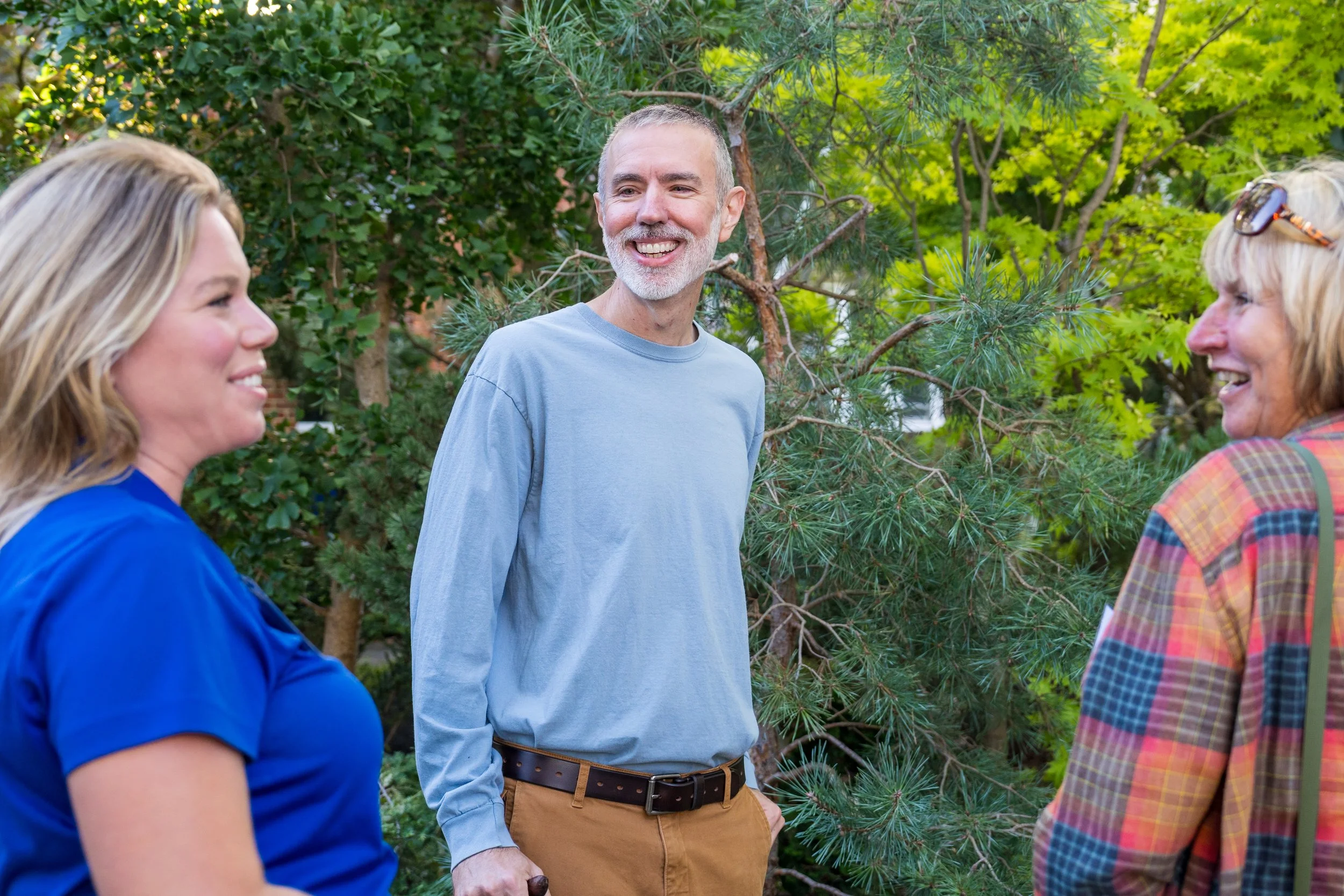 Three adults smiling and chatting outdoors in front of green trees.