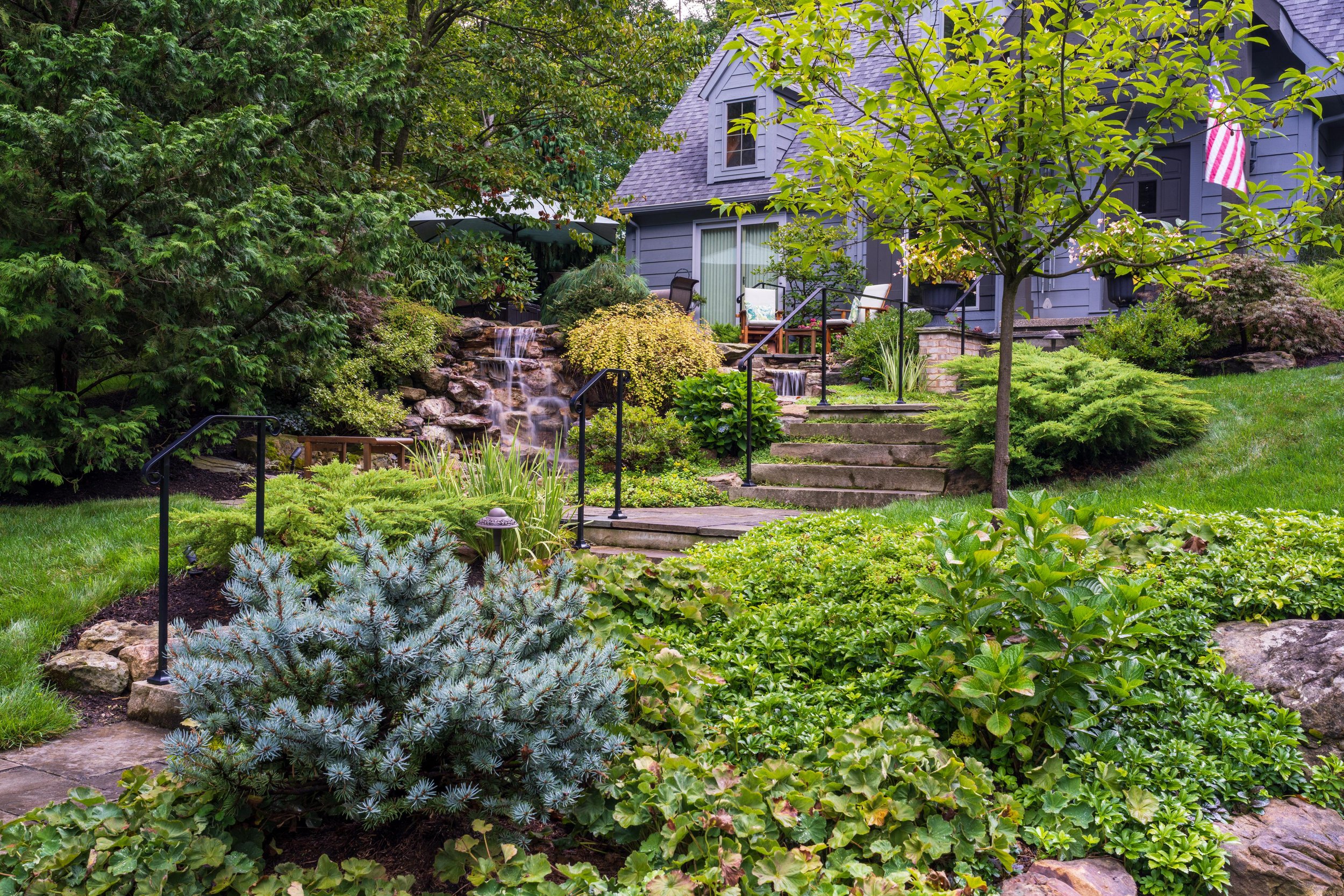 A landscaped backyard garden with a stone waterfall feature, green plants, a small tree, and outdoor seating in front of a blue house.