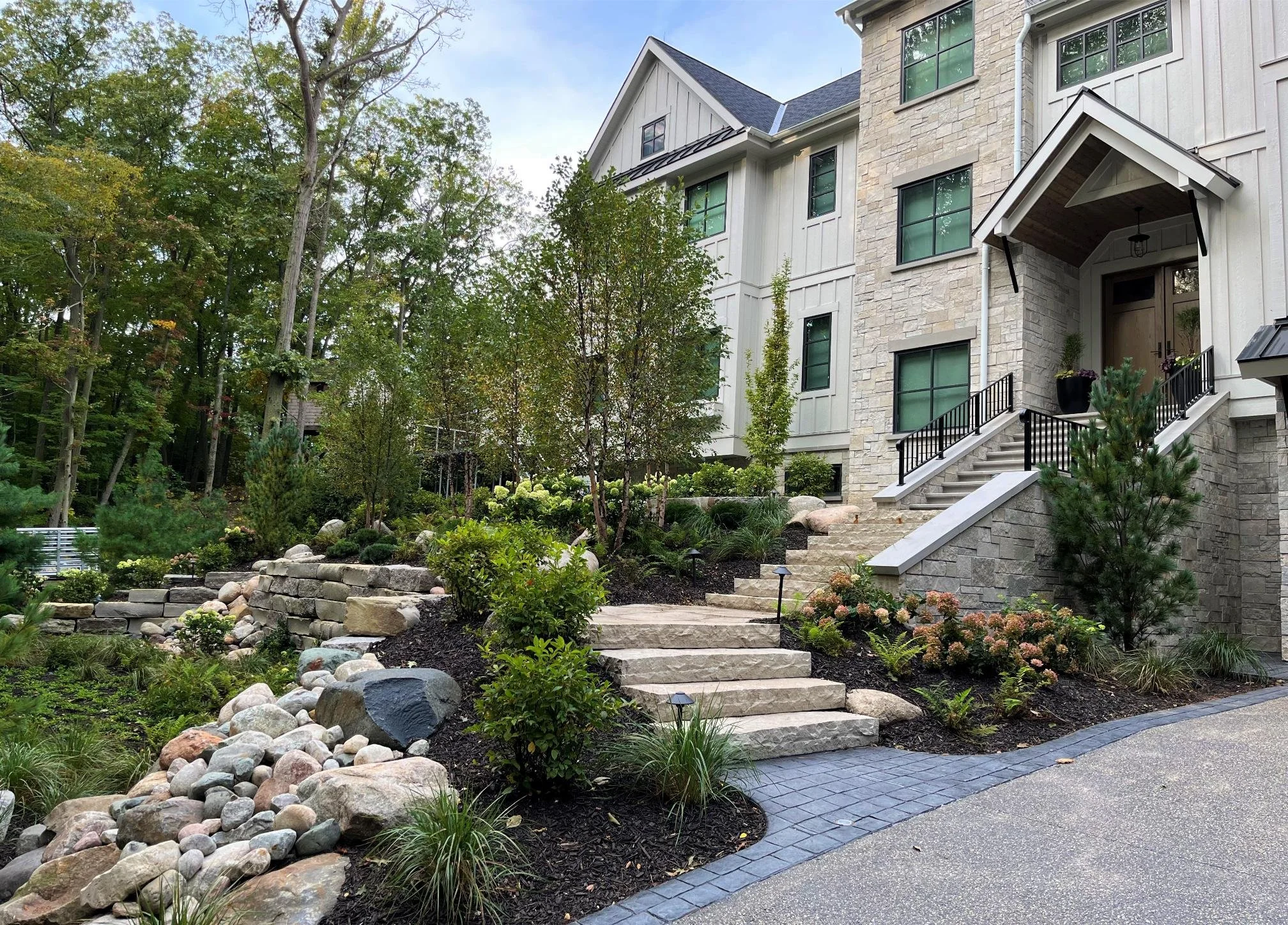A landscaped front yard featuring a stone pathway, stairs with black railings, various plants, bushes, and trees, with a modern multi-story house with stone and siding exterior in the background.