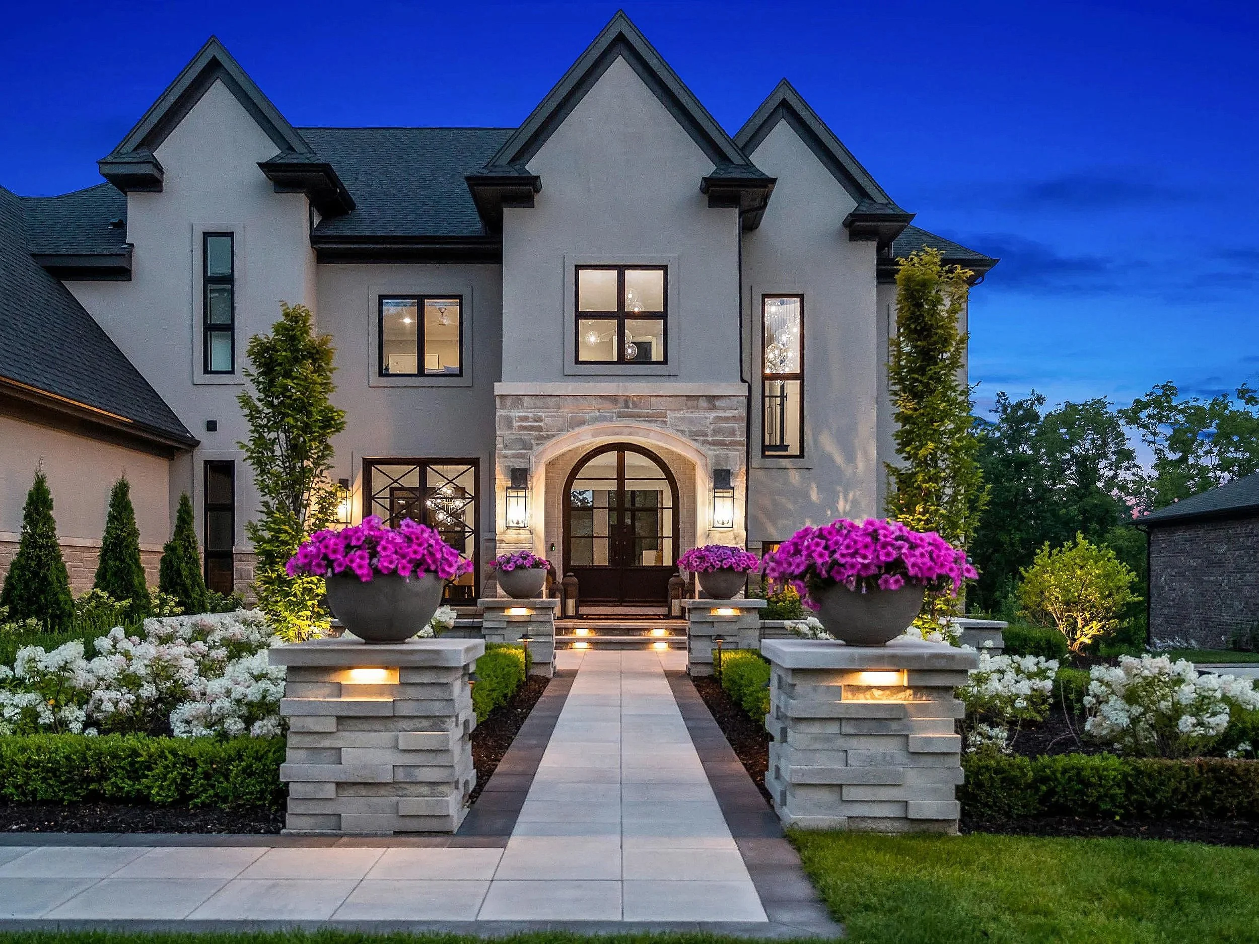 A large, modern house with a stone and stucco exterior, illuminated by exterior lights at dusk, featuring a landscaped front yard with pink and white flowers and a walkway leading to the entrance.