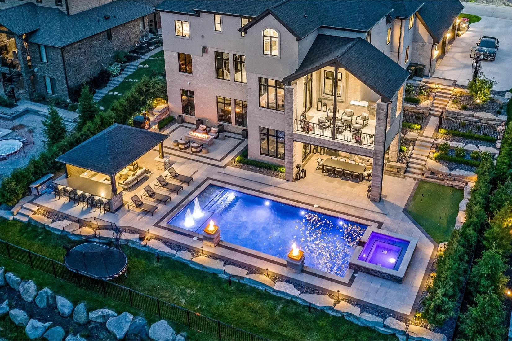 Aerial night view of a modern house with lit indoor and outdoor spaces, a swimming pool with fountains, lounge areas, a hot tub, and landscaped yard.