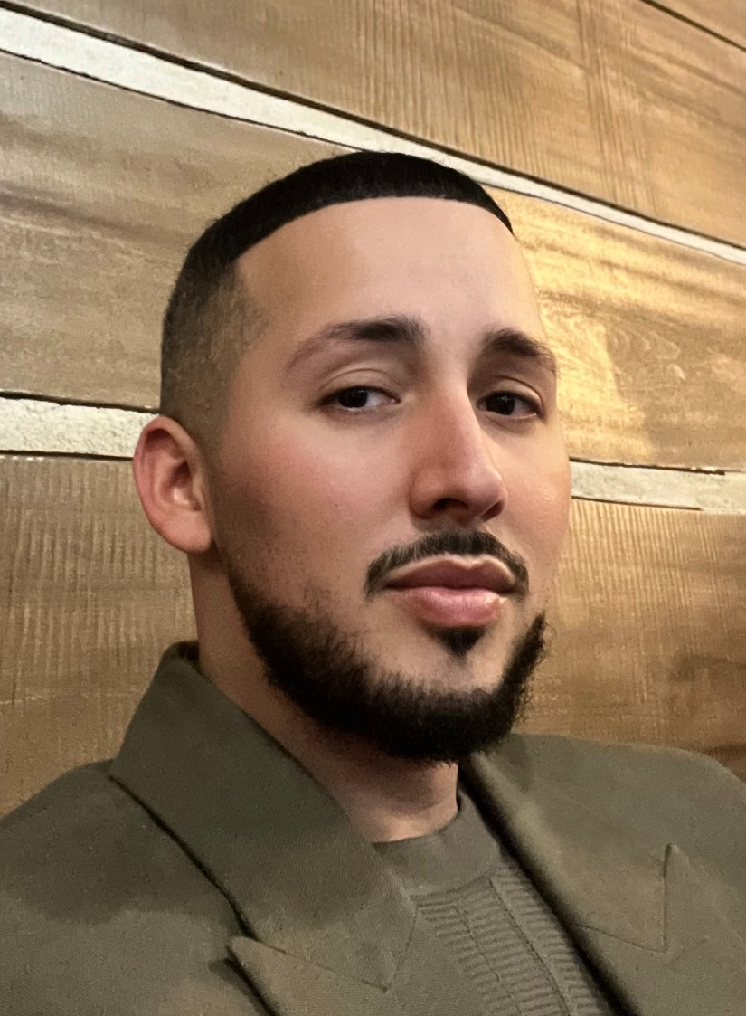 A young man with short dark hair and a beard, wearing a green jacket, sitting against a wooden wall.