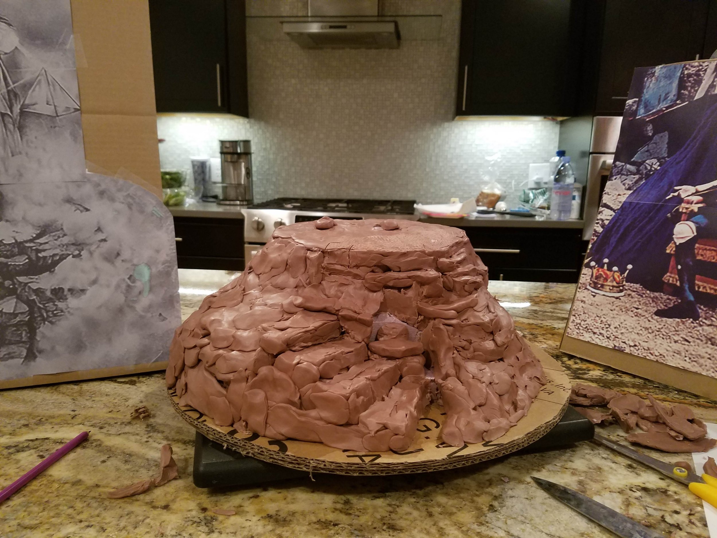 Unfinished clay sculpture of a turtle on a round base on a kitchen counter, with various art supplies and reference photos in the background.