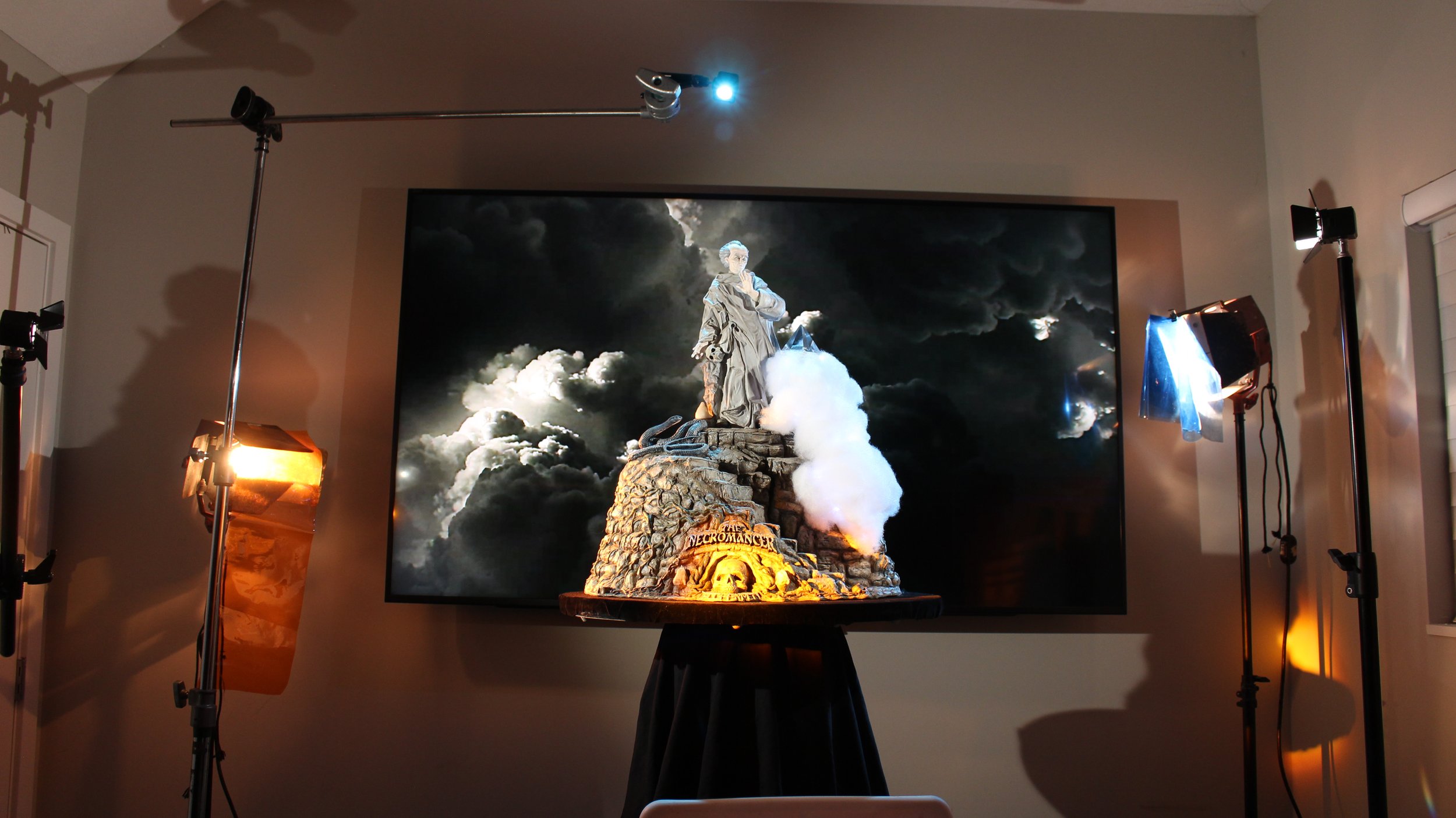 A detailed tabletop sculpture of a wizard monk standing on a rocky pedestal with the words 'Necromancer' engraved at the front, set against a large dark cloudy backdrop with a bright glowing moon. The scene is lit by professional studio lights around the sculpture.