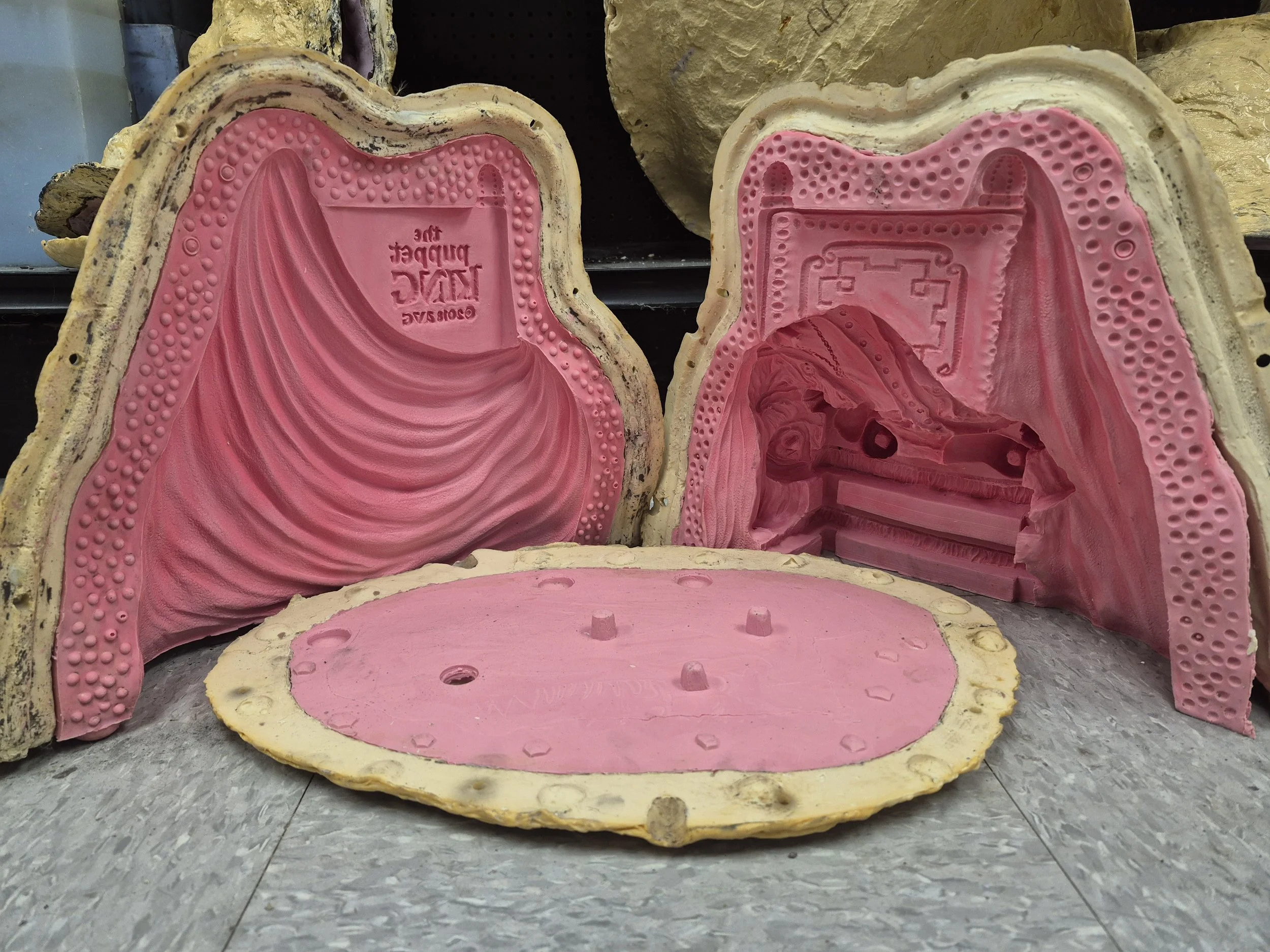 The image shows a cross-section of a pink and yellow silicone mold of a face, showing the detailed interior including eyes, nose, mouth, and other facial features, with the mold laying face up on a speckled floor.