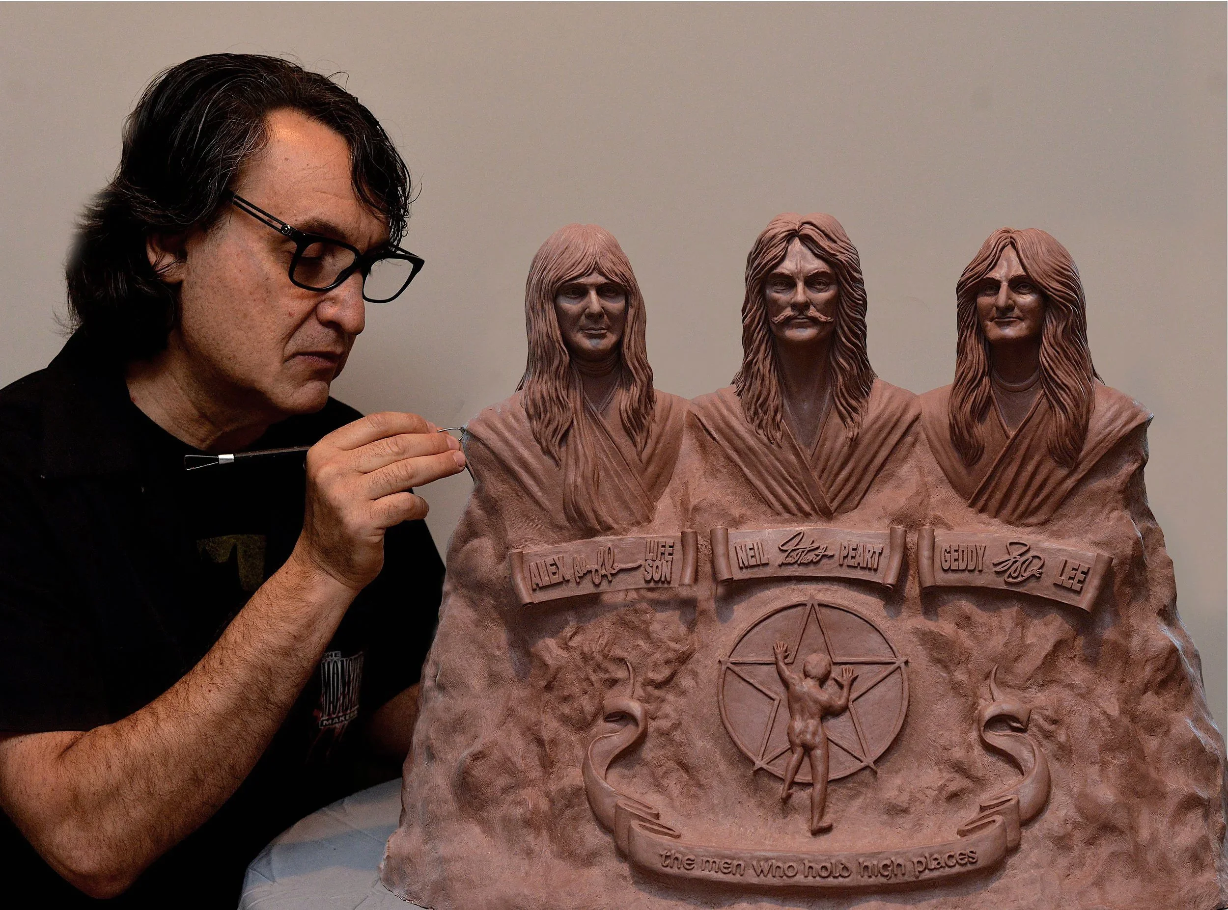 A man wearing glasses working on a clay sculpture of three figures. The sculpture has inscriptions and symbols at the bottom.