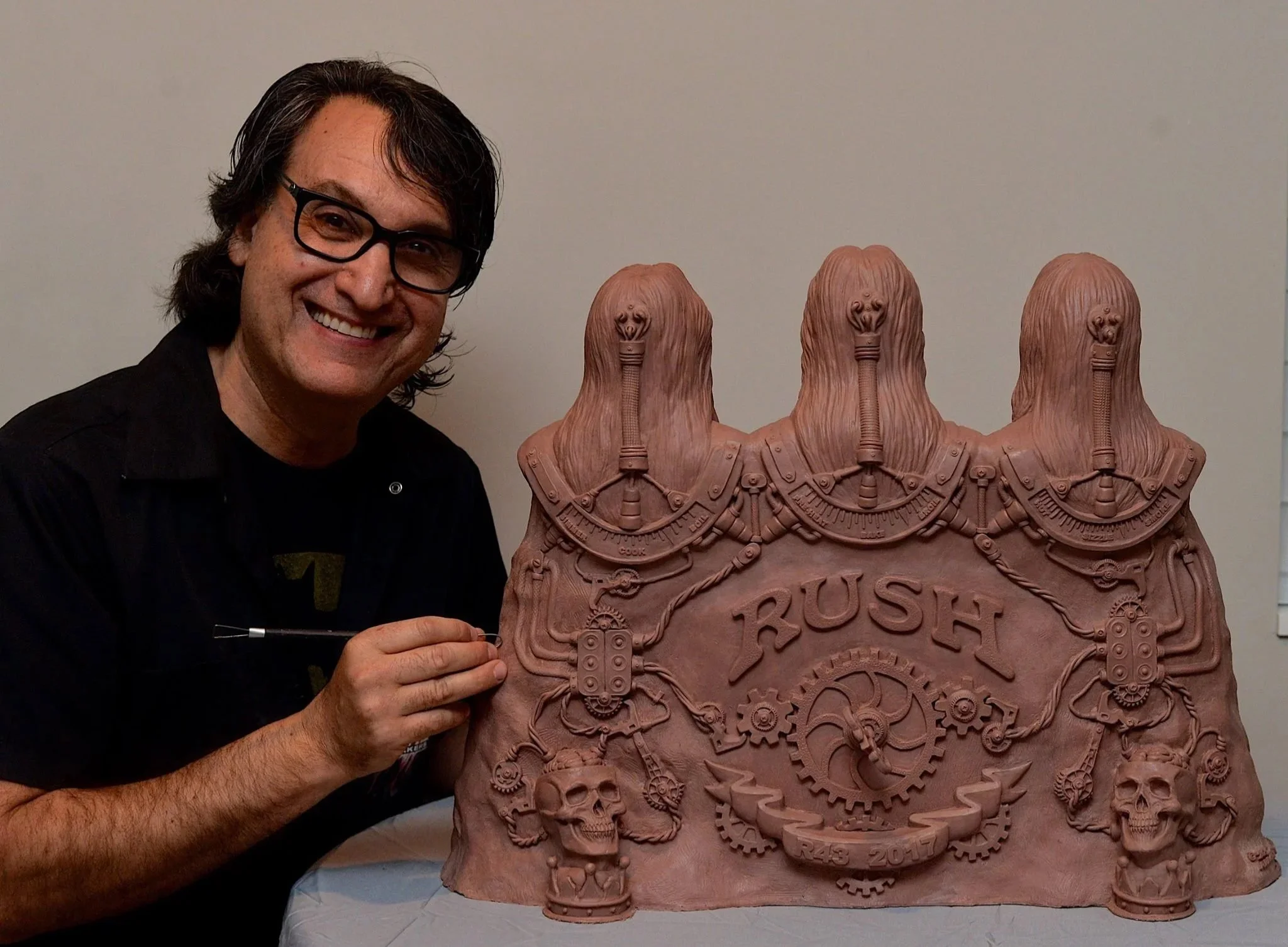 A man with glasses smiling and holding a small tool, standing next to a large, detailed sculpture of multiple heads and gears with the words 'RUSH 2017' at the bottom.