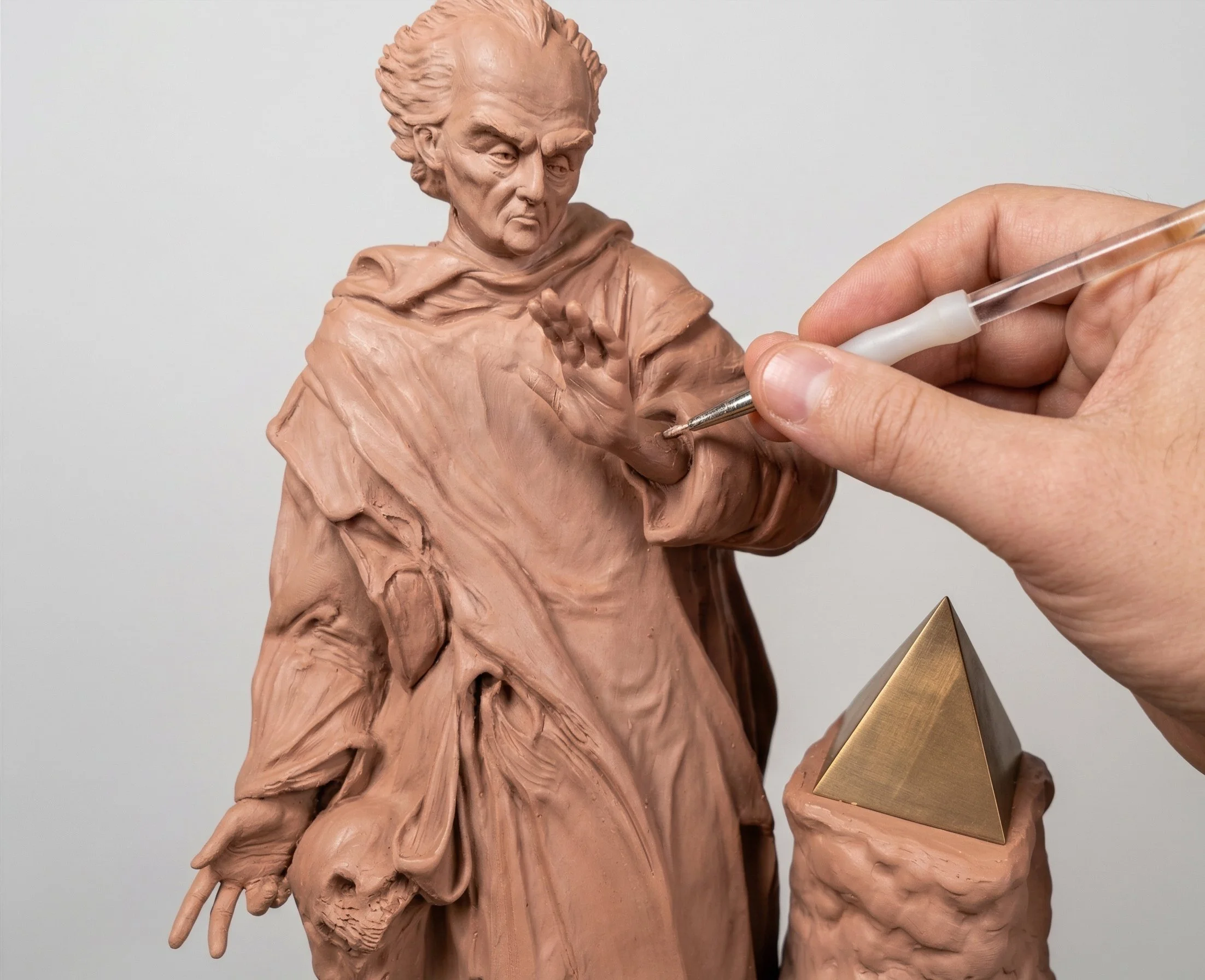 Close-up of a person sculpting a bust of an elderly man with a stern expression, with detailed facial features and wearing a draped garment; the sculptor's hand is holding a small tool, and a golden pyramid-shaped object is partially visible in the lower right corner.