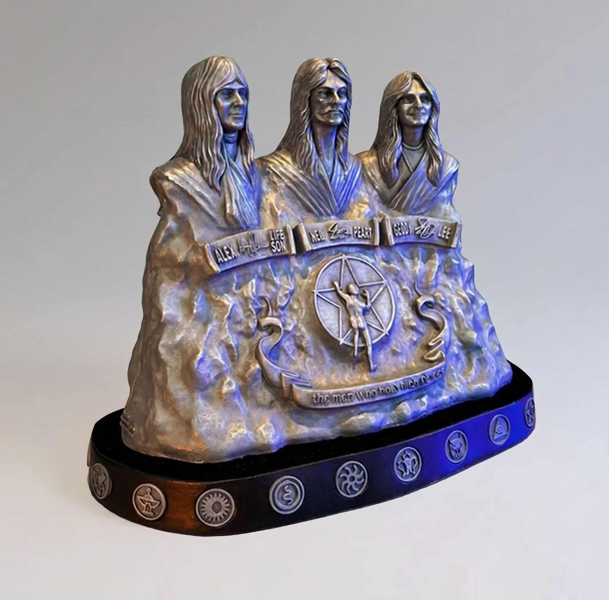 A metallic sculpture of three religious figures with long hair and robes, each labeled with names including Alex, Son, New, Peak, God, and Lee, with symbols inscribed on the base.