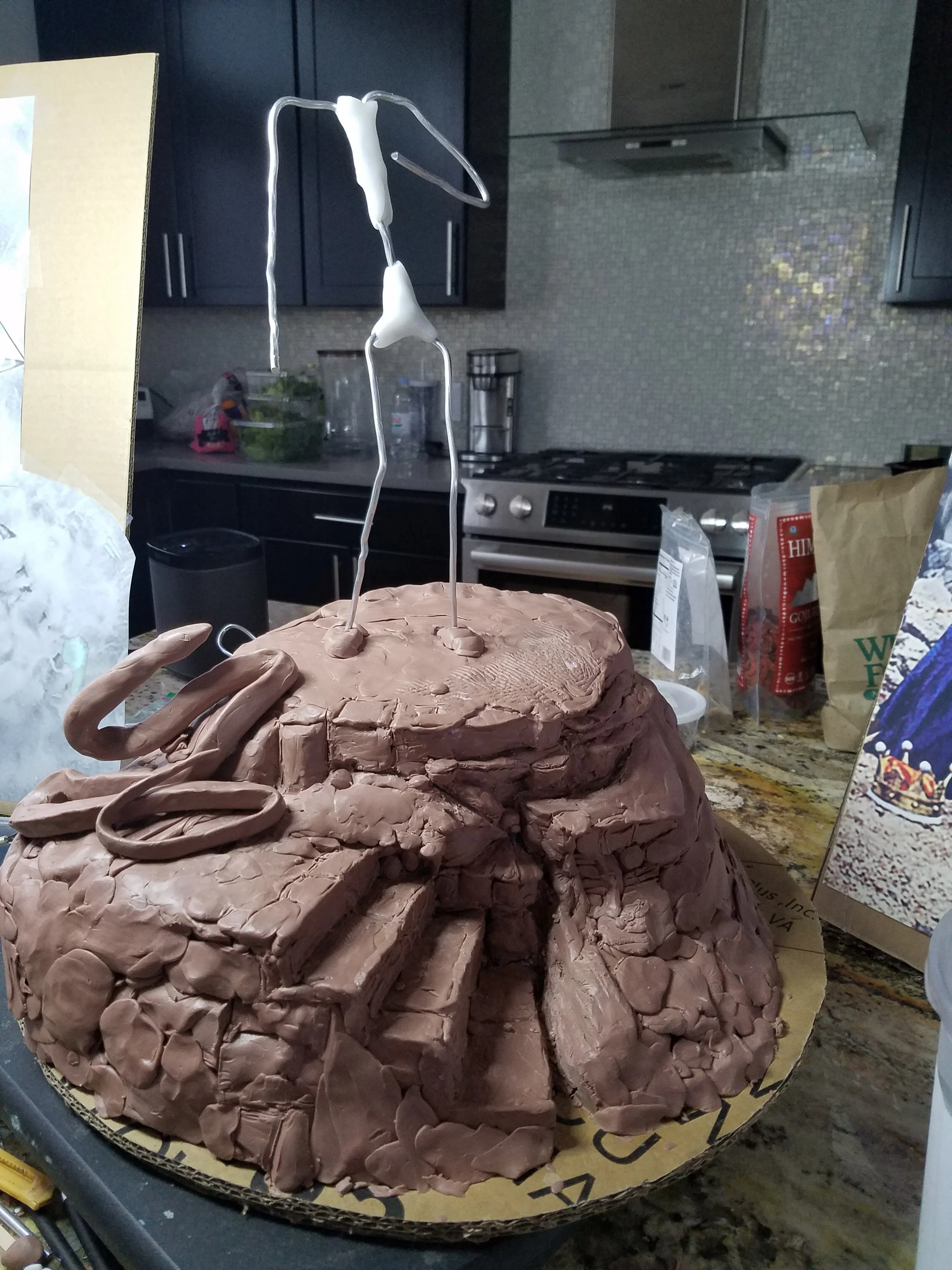 A cake made of brown modeling chocolate shaped like a sleeping dog with a small white hat on its head, placed on a round cardboard base. In the background, kitchen cabinets, a stove, and various kitchen items can be seen.