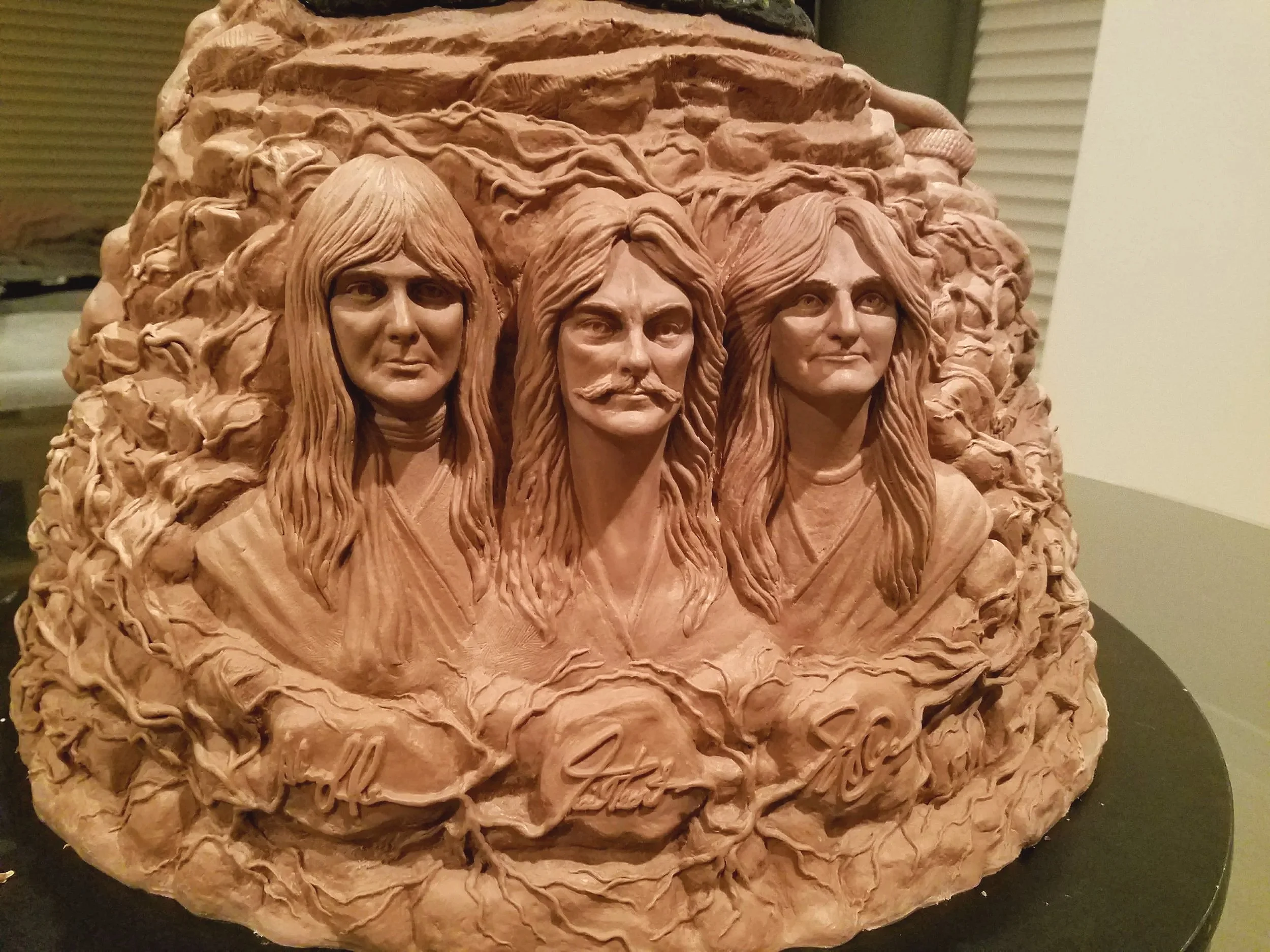 Terracotta sculpture of three faces with long hair, set against a textured, swirling background.