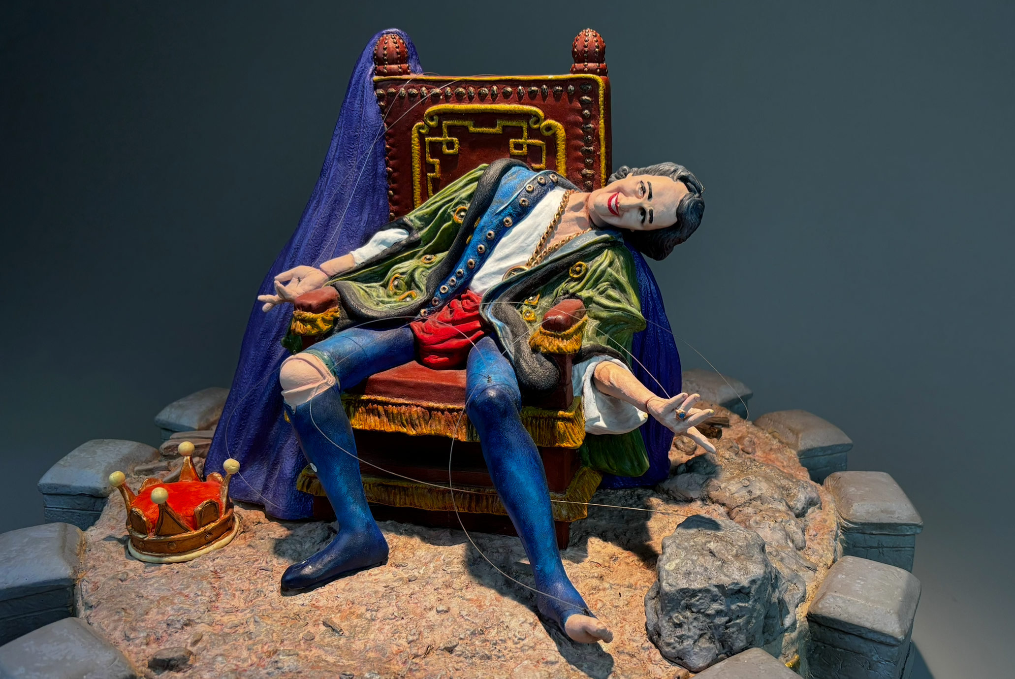 Colorful sculpture of a reclining man with long dark hair, dressed in elaborate clothing, sitting on a throne, with a crown on the ground nearby.