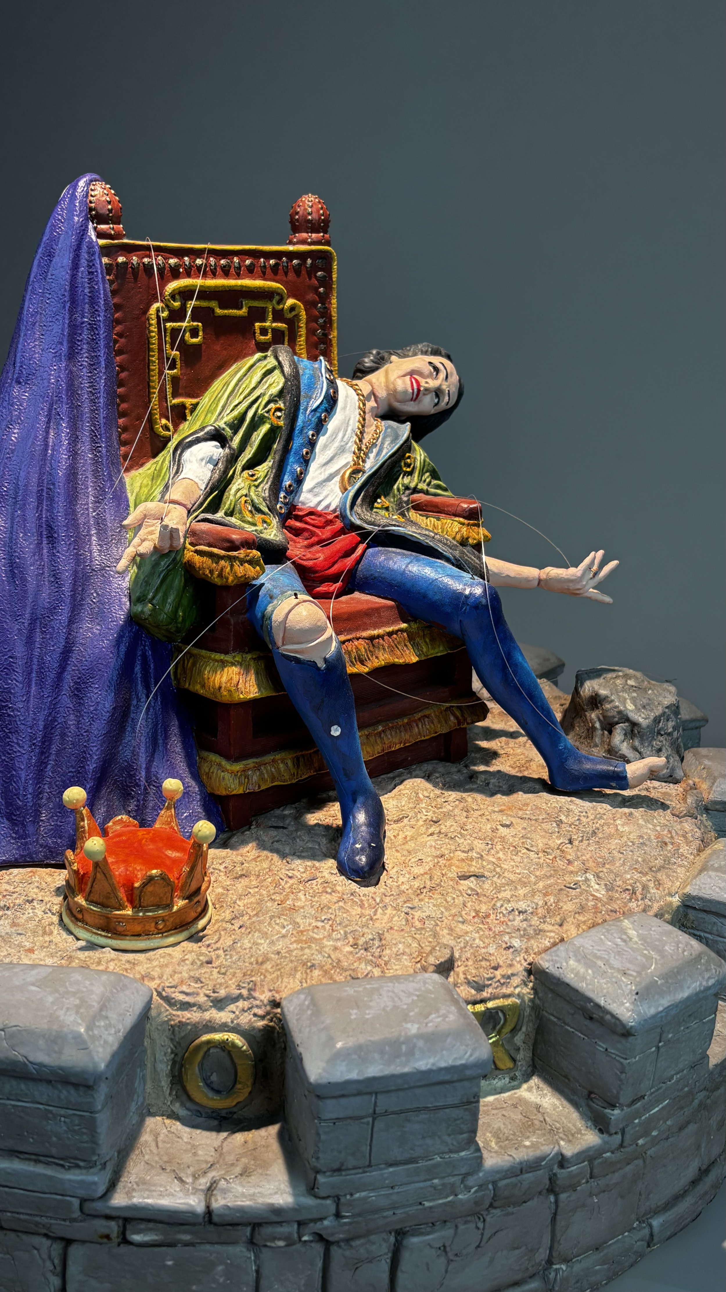 Colorful sculpture of a king reclining on a throne, with a purple drape on the side and a crown in the foreground, set on a rocky base.