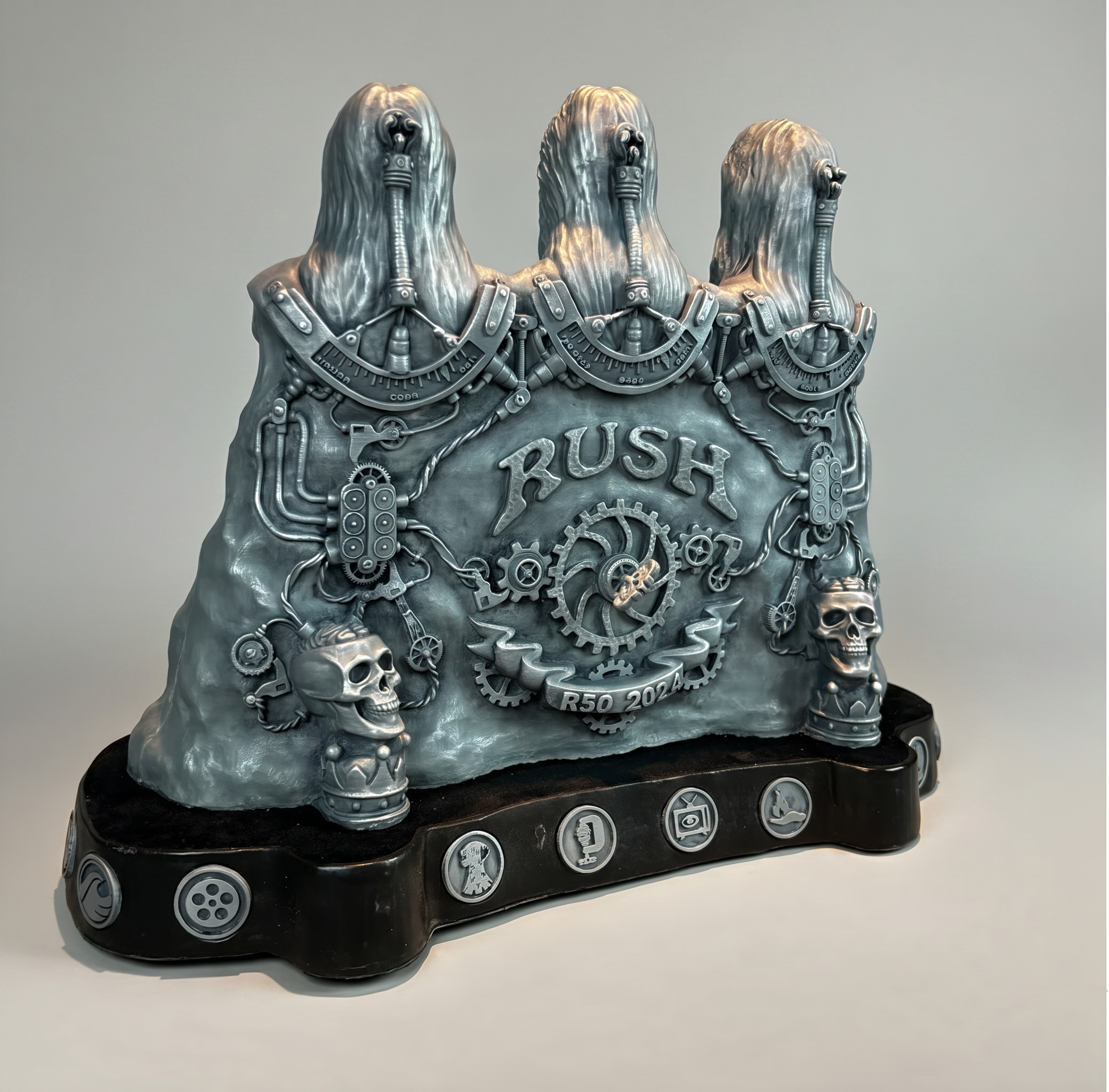 A metallic sculpture of three ghost-like figures with long hair, made of clock gears, cogs, and mechanical parts, with the word 'RUSH' inscribed on the front, and other mechanical elements and gears at the base.