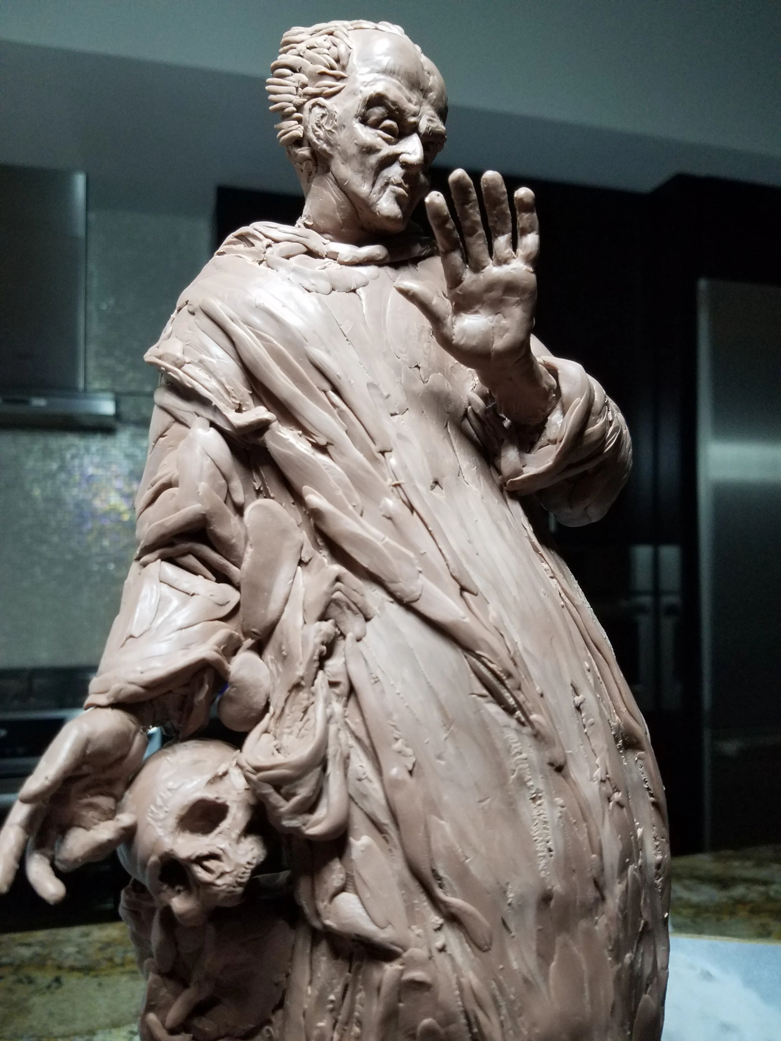 Close-up of the statue of a man with slicked-back hair, textured clothing with folds and details, hand raised in a gesture, set against a dark background.