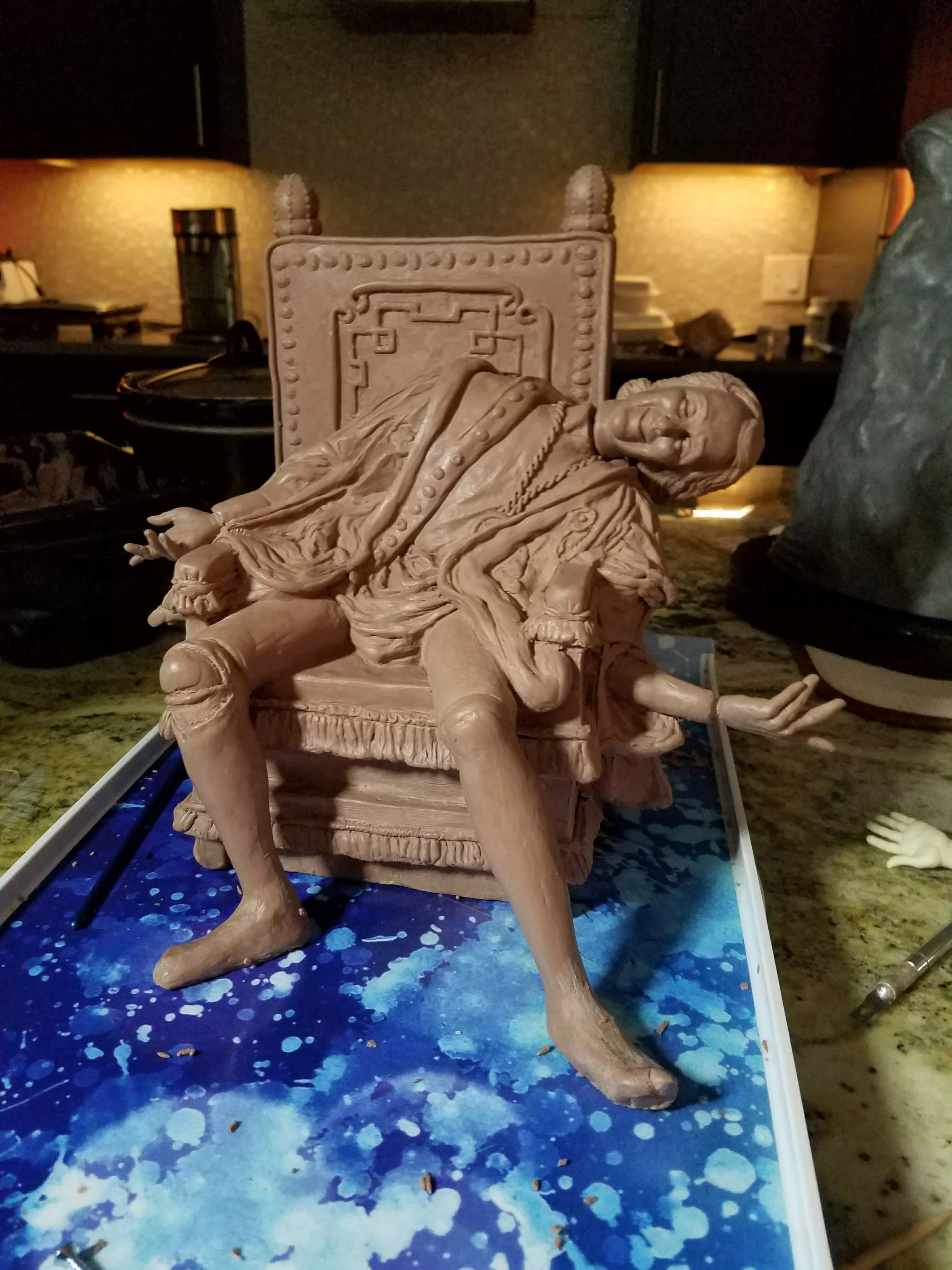 A detailed clay sculpture of a reclining woman on an ornate chair with a high back, situated on an artist's workspace with paint splatters on a blue surface. The background features warm lighting, a textured wall, and various art supplies.