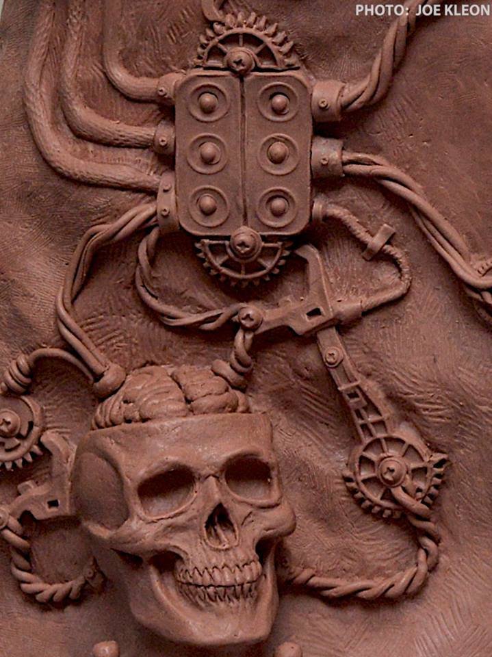 A detailed bas-relief sculpture featuring a human skull connected to various mechanical gears and wires, with a rectangular electronic device at the top.
