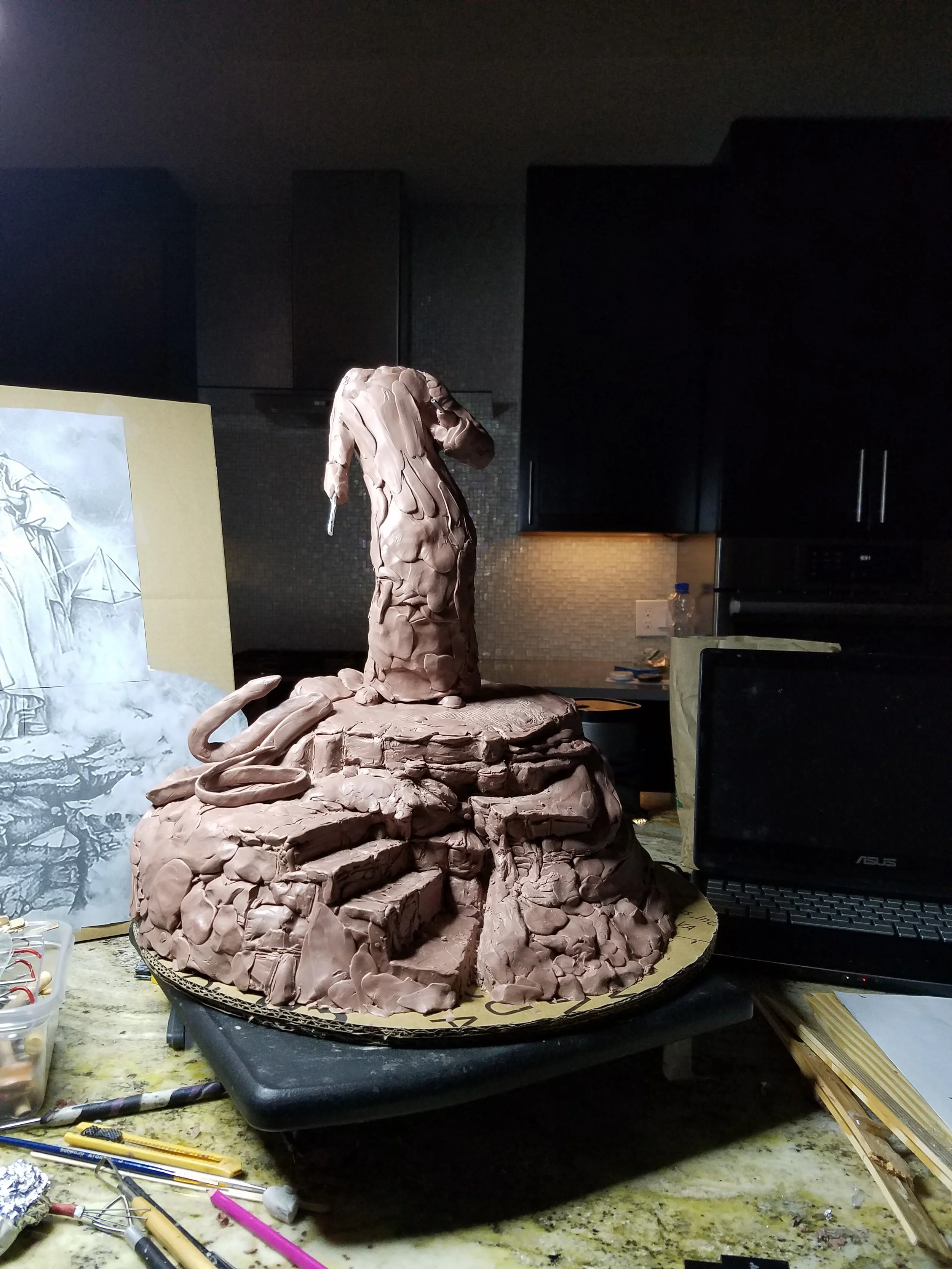 A clay sculpture of a snake coiled around a rocky base with a human head emerging from it. The sculpture is on a work surface in a workshop, with various tools and a partially visible computer nearby.