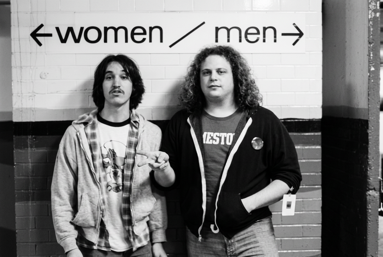 Two young men standing in front of a wall with a sign indicating directions to women and men restrooms. The man on the left has shoulder-length hair, a mustache, and is wearing a hoodie and a graphic T-shirt; he is pointing with his right hand. The man on the right has curly hair, is wearing a zip-up hoodie with a T-shirt underneath, and has a badge hanging from his pocket.