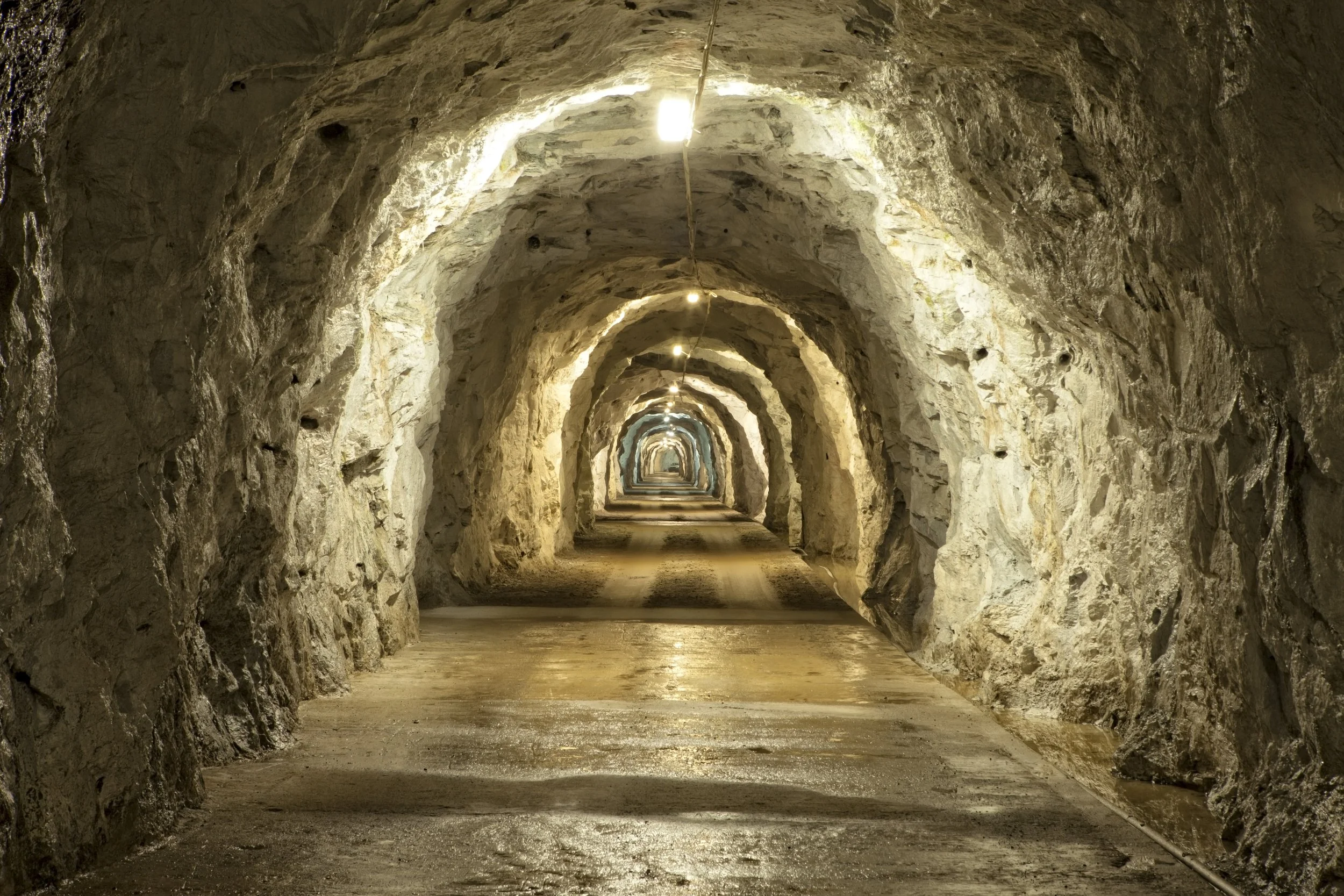 Underground tunnel with arched, rough stone walls and ceiling, lit by evenly spaced overhead lights, with a dirt floor.