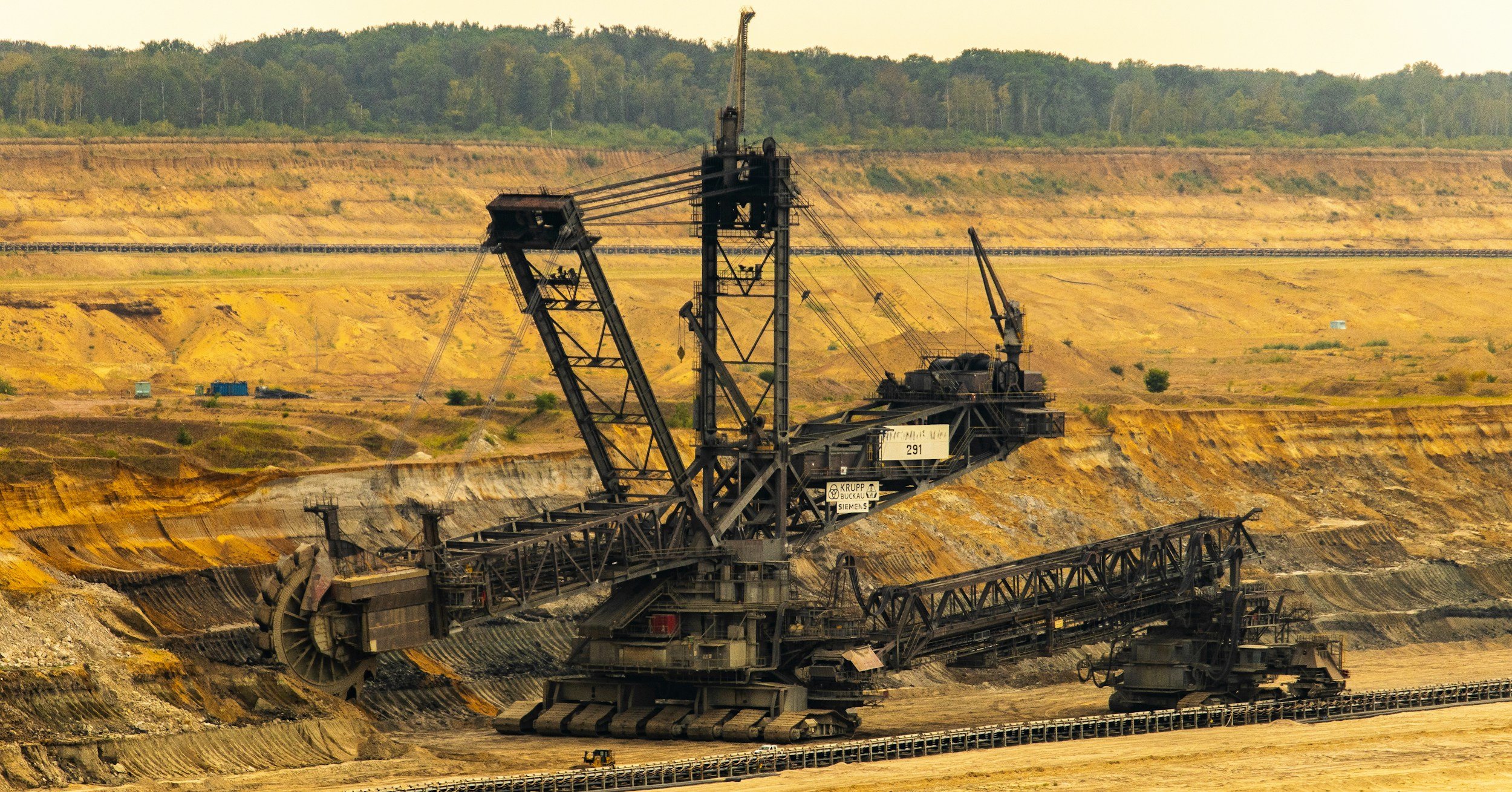 Large excavator working at a mining site with terraced earth and a forested background in the distance.