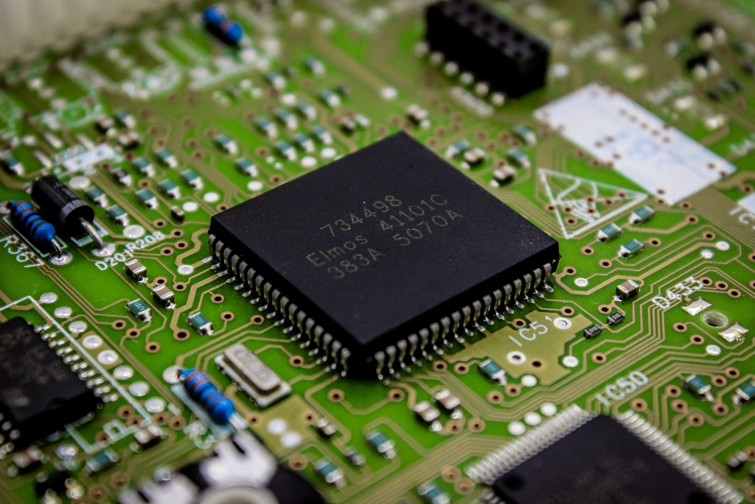Close-up of a green printed circuit board (PCB) with various electronic components, including a large black integrated circuit chip in the center, resistors, capacitors, and other small components.