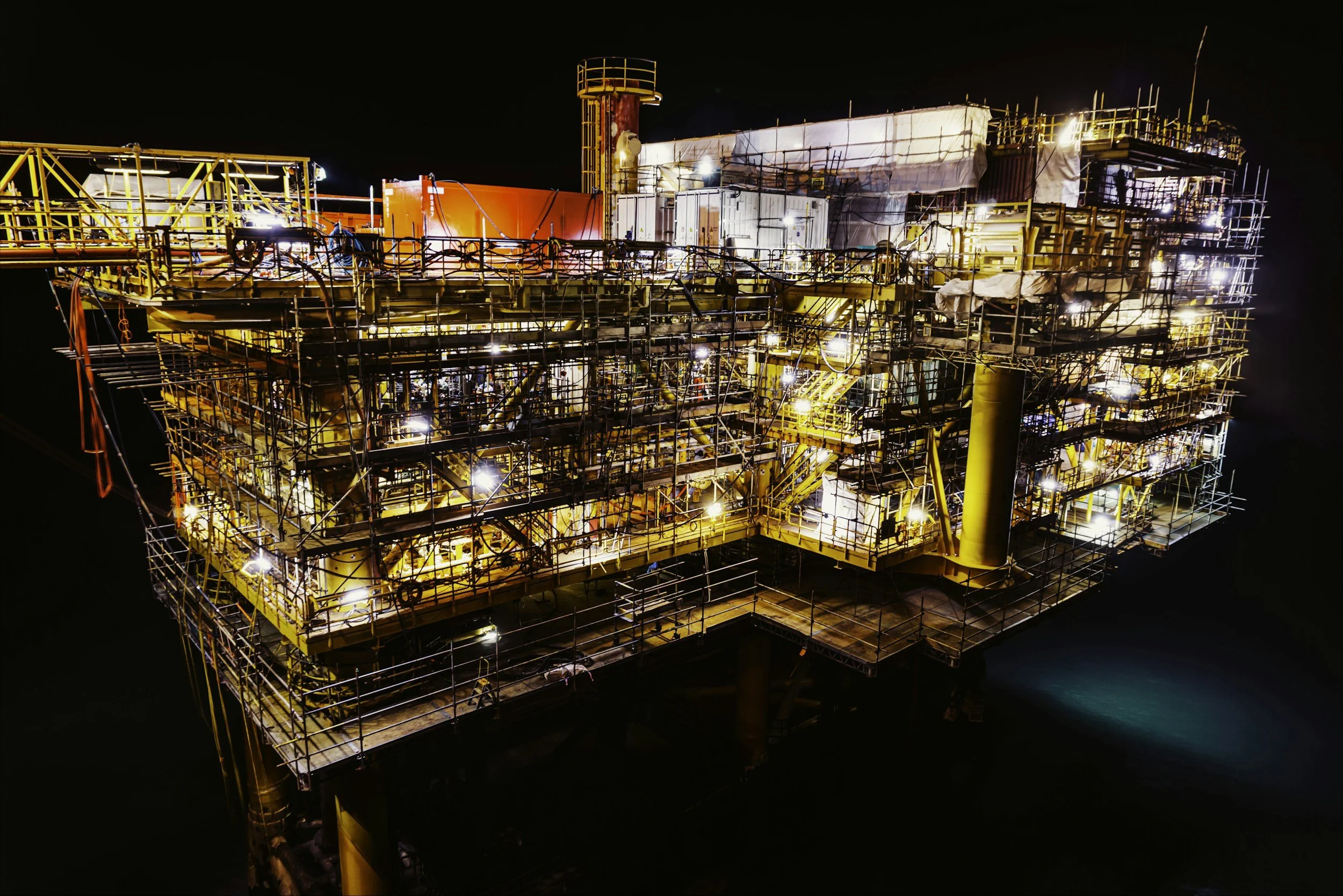 Offshore oil platform illuminated at night with complex scaffolding, pipes, and equipment visible against a dark sky.