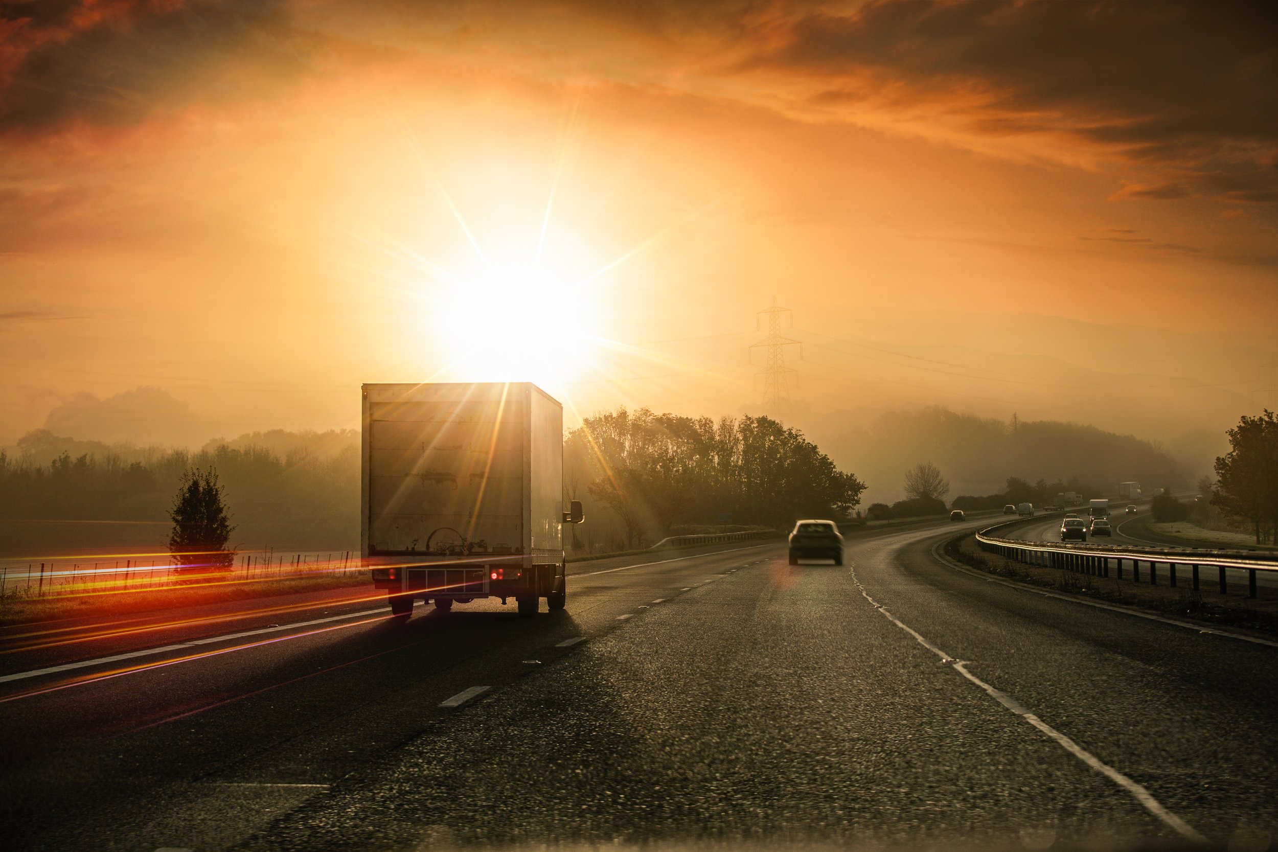 Sunset over a highway with cars and trucks, with a large truck in the foreground and power lines in the background.