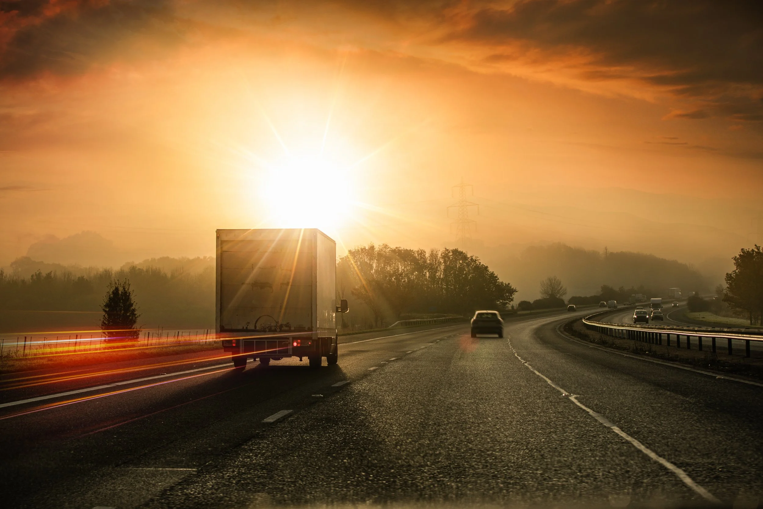 Sunset view from a highway with cars and trucks, with the sun near the horizon and reflecting light on the road, with some trees and utility poles in the background.