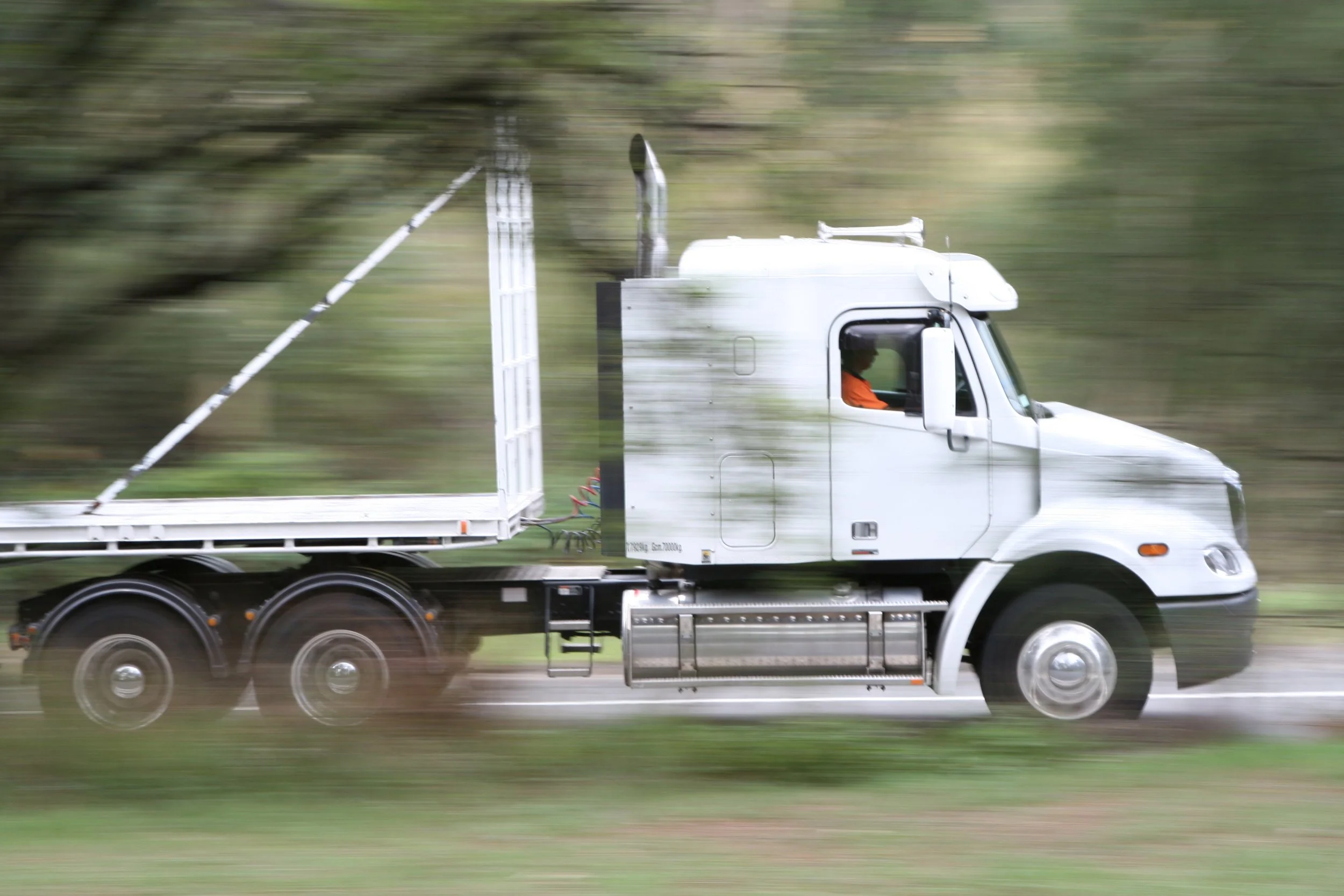 A white semi-truck in motion on a road surrounded by blurred greenery, with a tall load in the back and an orange-clad driver visible through the side window.