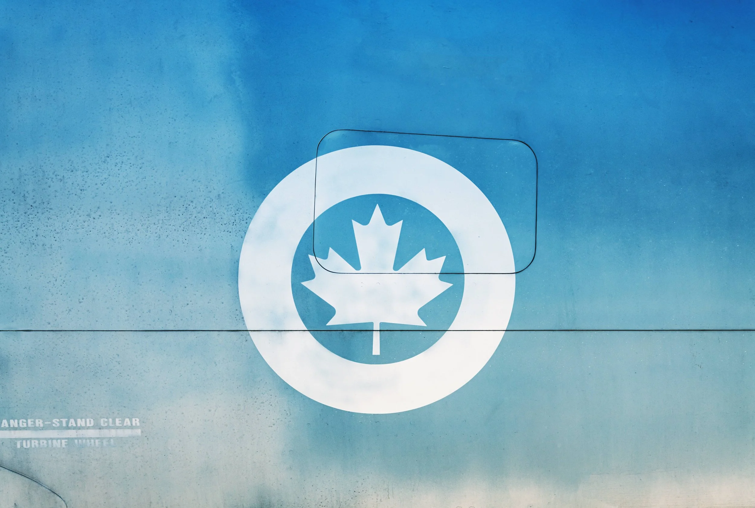 Close-up of a blue industrial panel with a white symbol of a maple leaf inside a circle and a warning label that reads 'DANGER - STAND CLEAR TURBINE WHEEL' at the bottom.