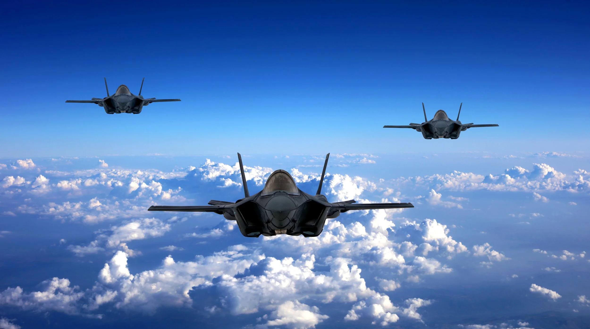 Three fighter jets flying in formation above the clouds against a bright blue sky.